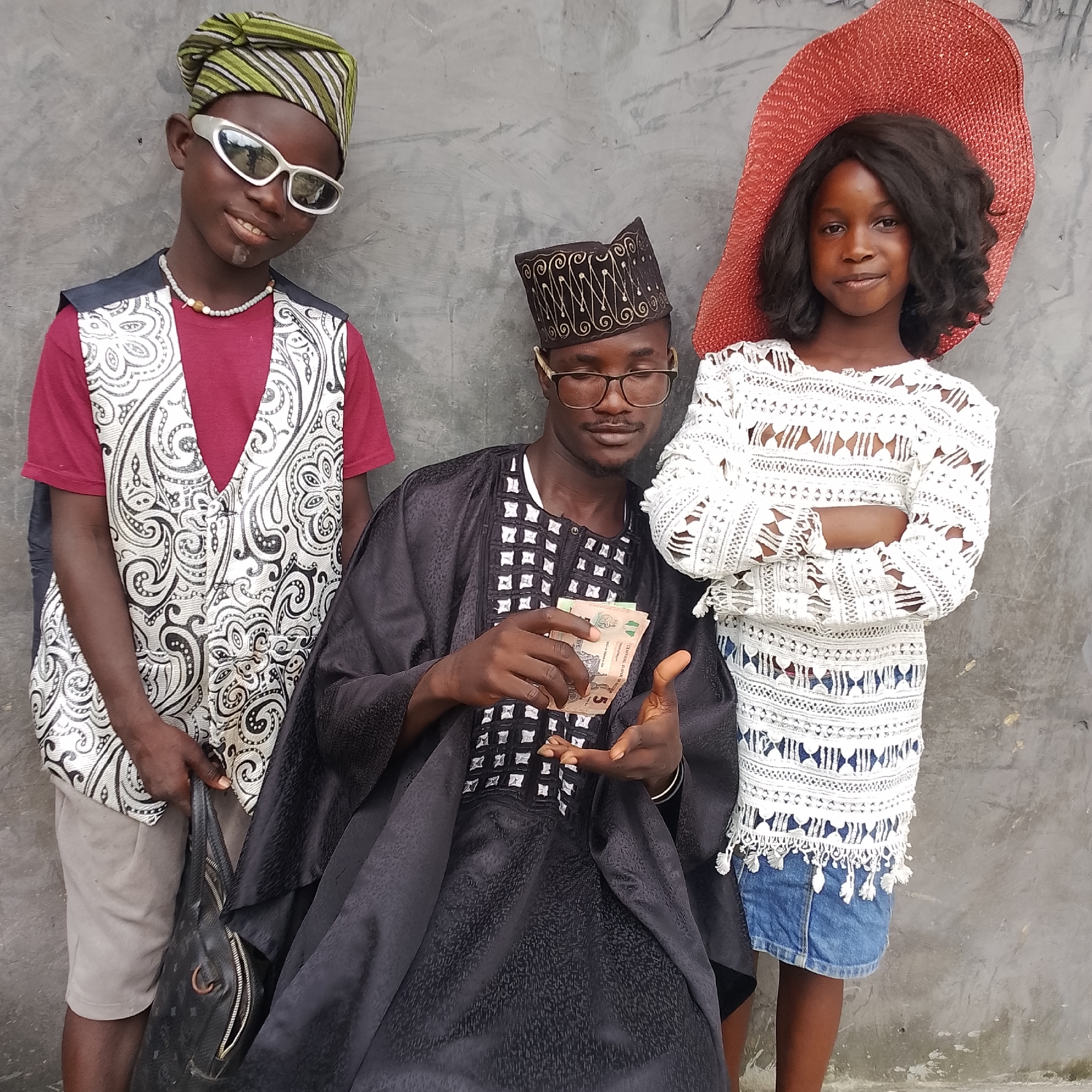 In Ado, Nigeria, three individuals stand against a grey, textured wall. On the left, a young boy with a green and striped cap and white sunglasses smiles, wearing a red shirt and a patterned vest. To his right, a man in a black, embroidered traditional outfit and glasses holds several banknotes, appearing to be counting them. On the far right, a young girl with a large red hat and a white, lacy top poses with her arms crossed. A black bag rests on the ground near the man. The scene appears to be outdoors in an urban setting.