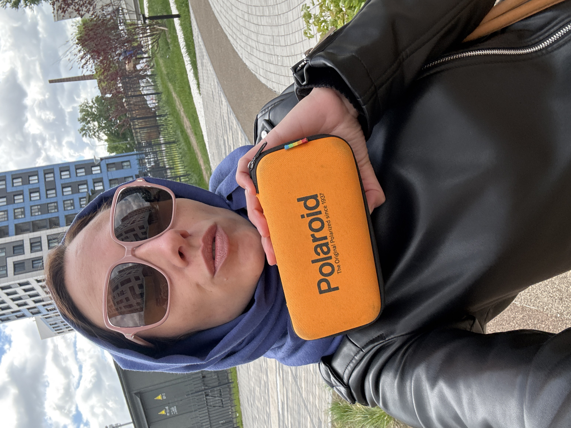 A person wearing pink sunglasses and a blue head covering is holding an orange Polaroid case in front of a city landscape. The person is wearing a black leather jacket. The background includes a modern residential building with multiple windows, trees, a fence, a paved walkway, and a cloudy sky. The location appears to be in Saint Petersburg, Russia, based on the architectural style and urban environment.