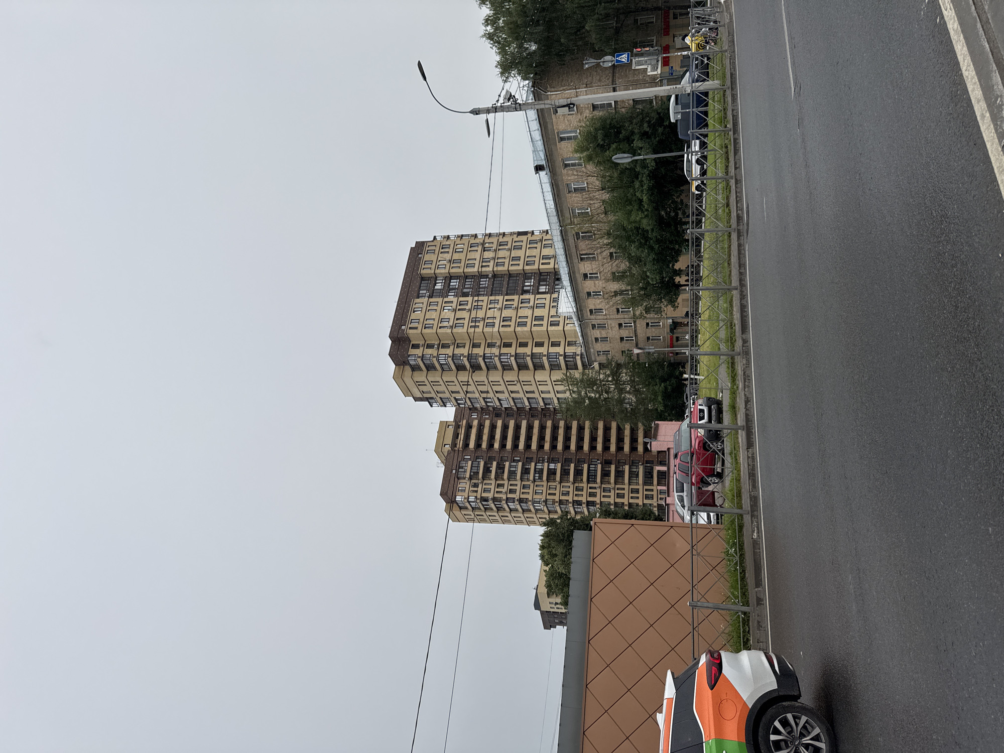 The image shows a street scene in Shchyolkovo, Russia, on an overcast day. In the foreground, a wet asphalt road is visible with a white line marking the edge. To the left, the rear of a white car with orange and green decals is parked. Behind the car, a section of a building with a brown, diamond-patterned facade is seen. Beyond this, a metal fence separates the sidewalk from a grassy area where several cars are parked, including a red SUV and several white and dark-colored vehicles. In the background, two tall apartment buildings with light-colored facades and numerous balconies dominate the skyline. To the right of the apartment buildings, a lower, older brick building with many windows stands. Trees are scattered throughout the scene, and a street lamp with multiple lights is positioned on the right side of the image. Pedestrians and cyclists are present on the far right, near a traffic signal.