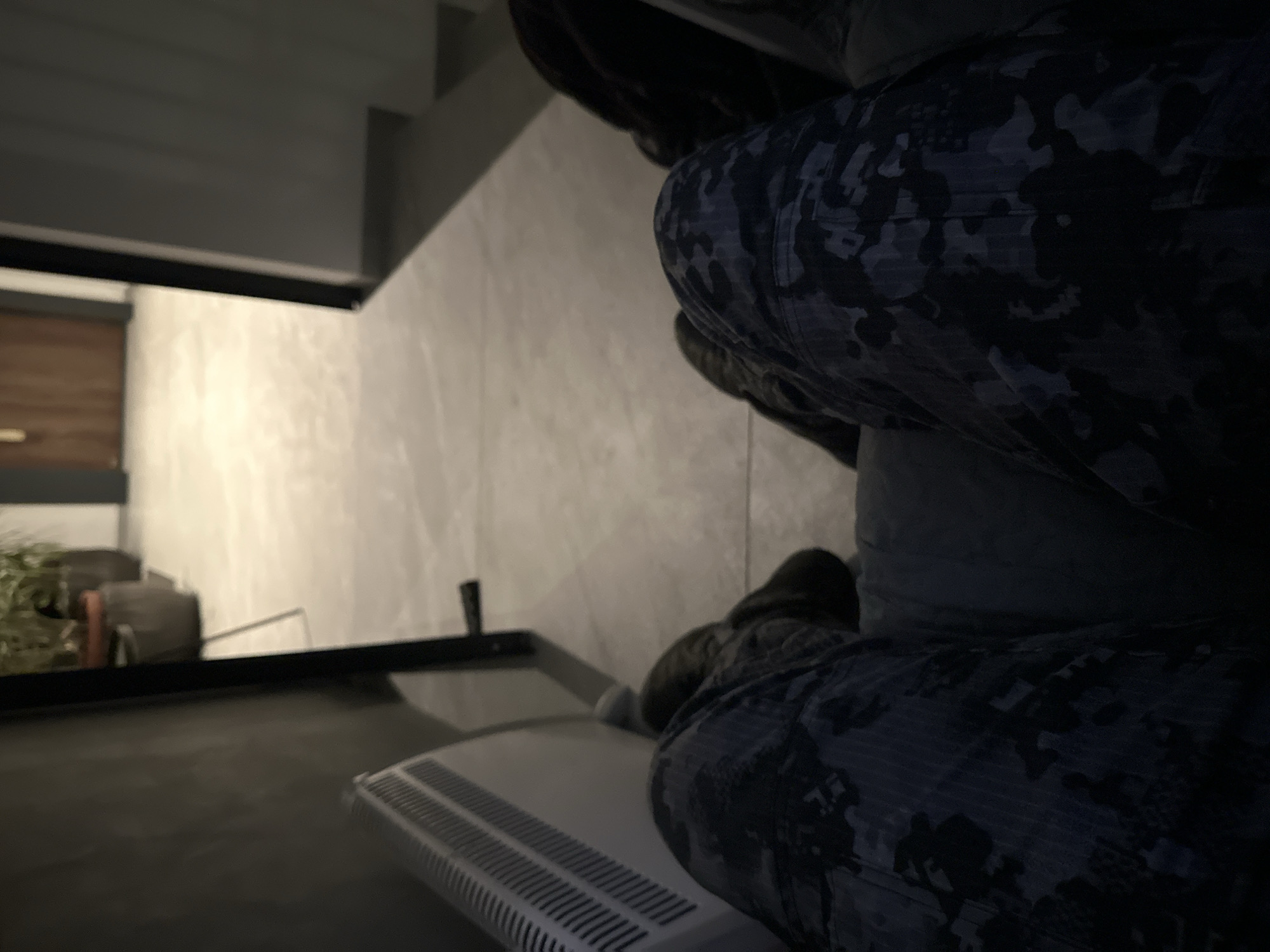 The image shows a person wearing camouflage pants sitting on a white radiator against a neutral-toned wall. A framed opening reveals a glimpse of an interior space with a wooden door and greenery. The location is Sofia, Bulgaria.