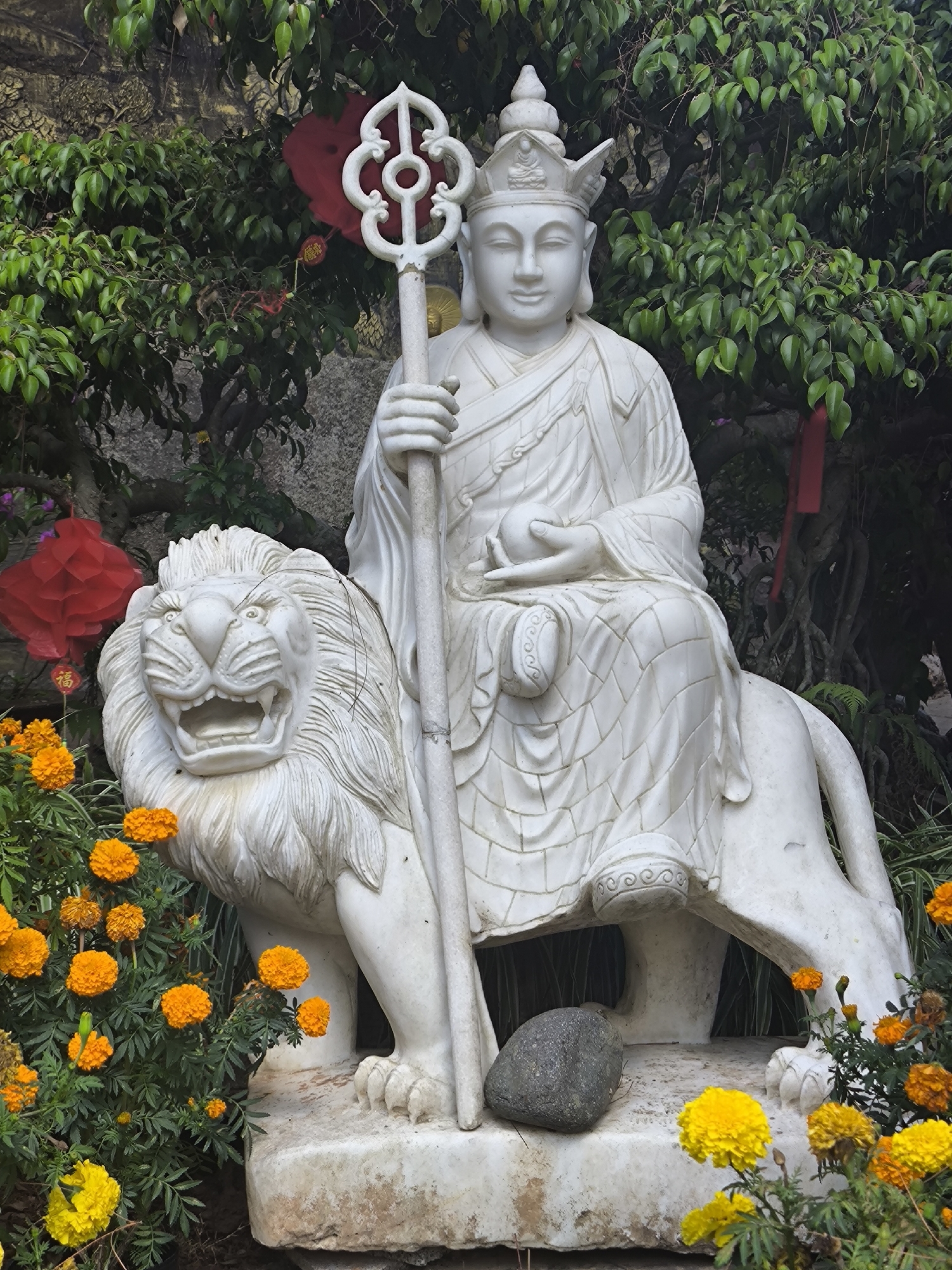 The image depicts a white marble statue of a Buddha-like figure seated atop a lion. The figure wears a crown-like headdress, has a serene expression, and holds a staff-like object with a circular design in one hand and what appears to be an orb in the other. The lion is depicted with an open mouth and a thick mane. The statue is surrounded by greenery and flowers, with orange and yellow flowers prominent in the foreground. Red paper lanterns are visible in the background. The scene is located in the city of Lâm Viên - Đà Lạt, Vietnam.