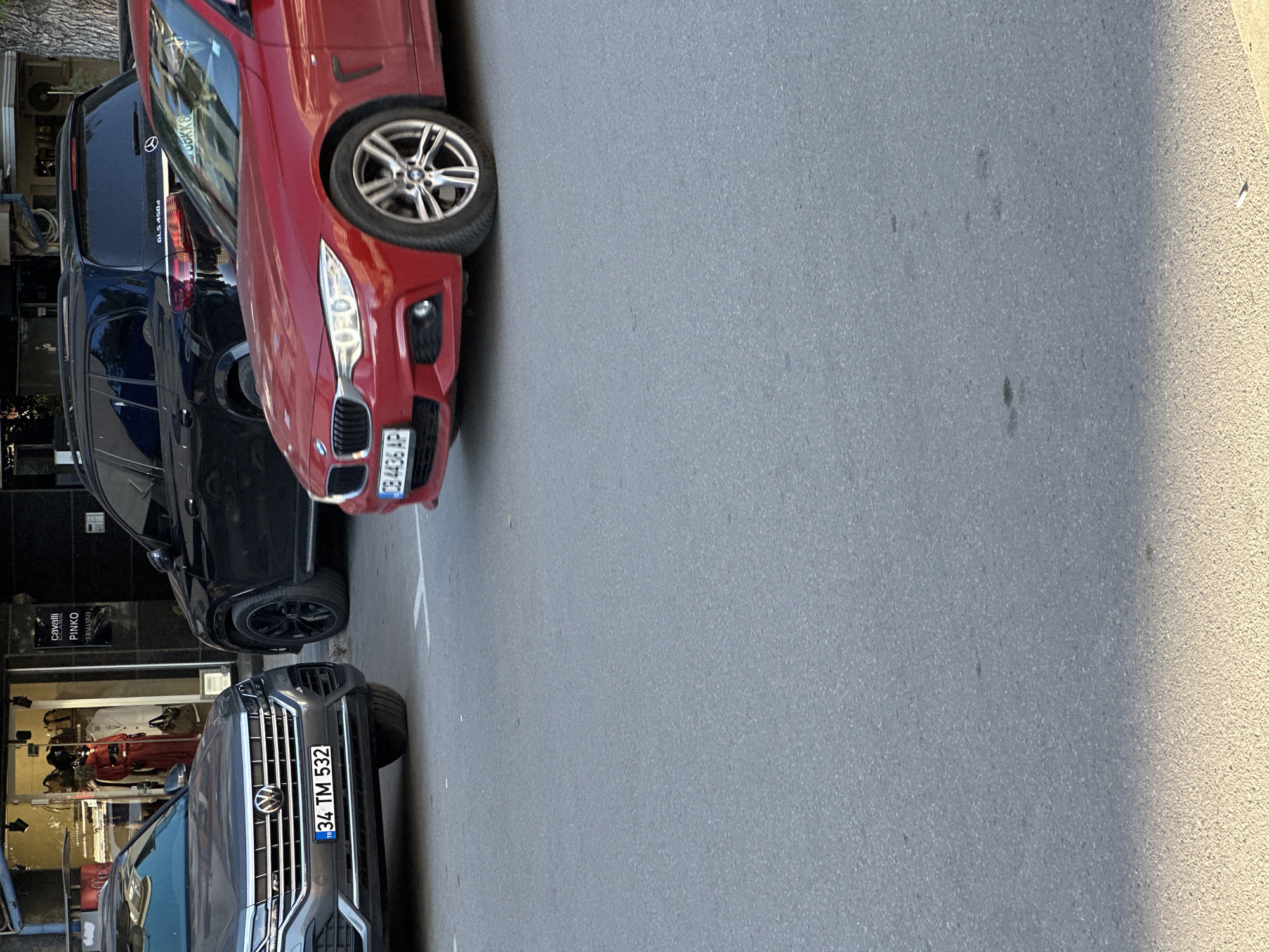 The image shows a street scene with several parked cars. The cars are a red BMW, a black Mercedes-Benz GLS 450d, and a gray Volkswagen. The license plate of the BMW is "GB 4436 AP," and the license plate of the Volkswagen is "34 TM 532." The cars are parked on the side of the street. Behind the cars, there is a store with mannequins dressed in red clothing. The store's name is partially visible, showing "Pinko" and "Cavalli Germano."
The image was most likely taken in Turkey because of the Turkish license plate.