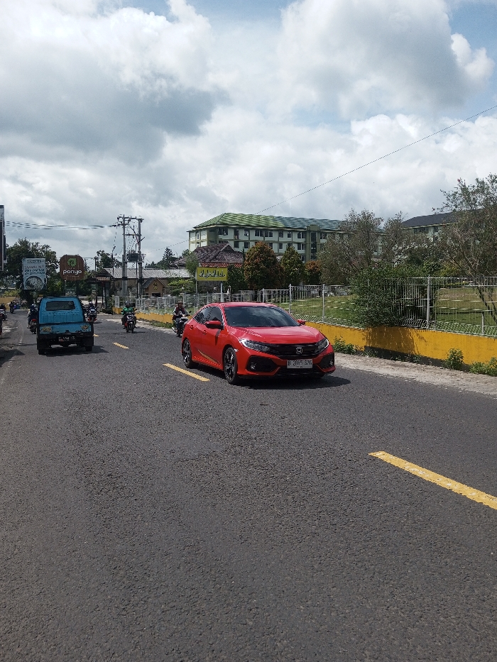 The scene depicts a daytime street view featuring vehicular traffic under an overcast sky. A red Honda Civic sedan, displaying Indonesian license plate "B 201 F 57G," occupies the right side of the asphalt road. To its left, a light blue pickup truck, an older model, moves in the same general direction. Several motorcycles, some with riders, are also visible on the road. Yellow dashed lines mark the lanes. On the right, a yellow concrete barrier separates the road from a grassy area secured by a metal fence. In the background, a large multi-story building with a light green roof and light-colored walls stands prominently. Further left in the background, power poles, smaller buildings, and commercial signage, including a green sign bearing the text "ponyo," are present. The license plate "B 201 F 57G" identifies the vehicle as registered in Jakarta, Indonesia, which contradicts the provided location of Gubbisamamidi, India.