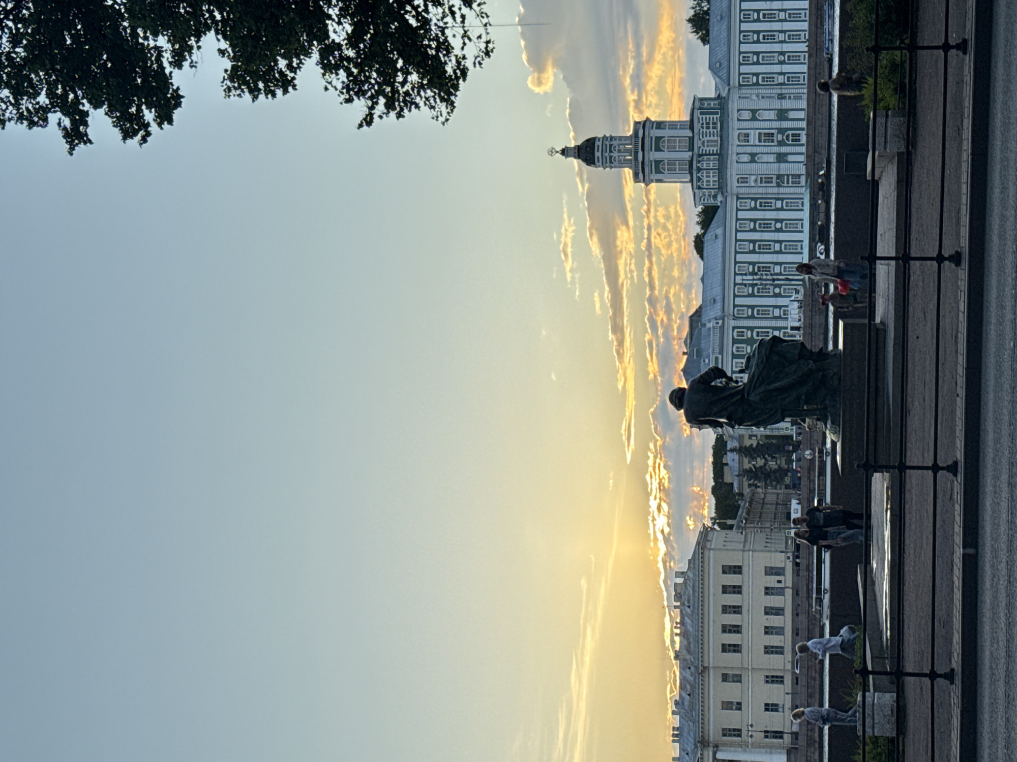The image depicts a sunset scene along the Neva River in Saint Petersburg, Russia, with the Kunstkamera building prominently visible. In the foreground, a dark bronze statue of a male figure stands on a pedestal, facing away from the viewer towards the river. A black metal fence runs across the bottom of the frame, separating the viewer from a paved walkway.
On the walkway, several individuals are present. To the left, two women stand near the river's edge, one in a white top and the other in a light-colored outfit. Further to the right, a couple stands together, and another woman with a red bag is visible. The river itself has a body of water, with a boat visible in the distance.
The Kunstkamera building is a large, pale green structure with white trim and numerous windows. It features a distinctive domed tower topped with a spire. The sky is a gradient of pale blue to yellow, with dramatic clouds illuminated by the setting sun. The branches of a large tree are visible in the upper right corner of the image, partially obscuring the sky. The overall atmosphere is serene, with the warm light of the sunset casting a soft glow over the architectural landmarks and the figures present.