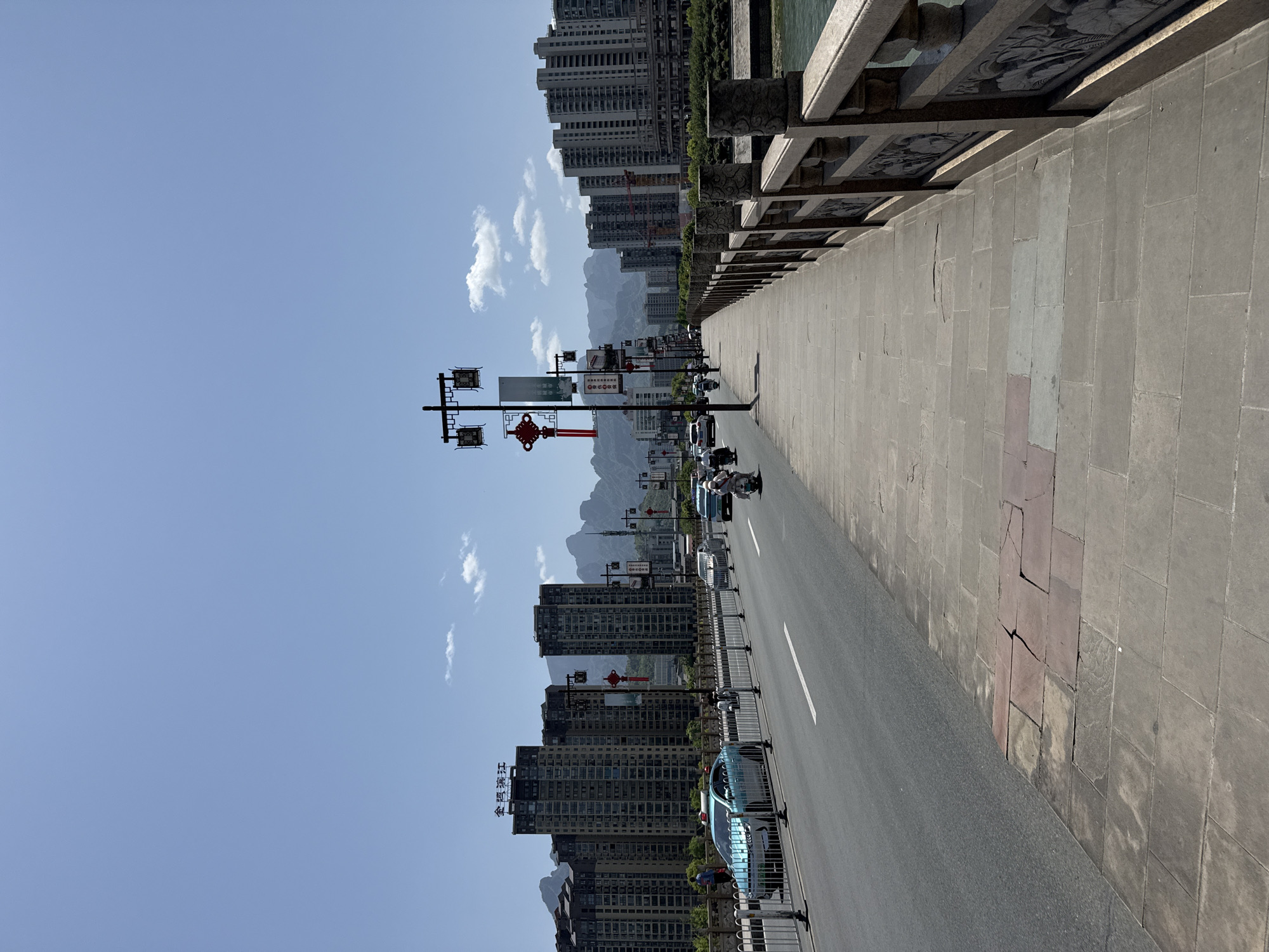 The image shows a view from a bridge in Zhangjiajie, China. The bridge has a stone walkway and a carved stone railing. A road runs parallel to the walkway, with cars and motorcycles visible. On the other side of the road, there are tall apartment buildings. Mountains are visible in the background, under a blue sky with scattered clouds. Decorative Chinese-style street lamps with red lanterns are also present along the road.