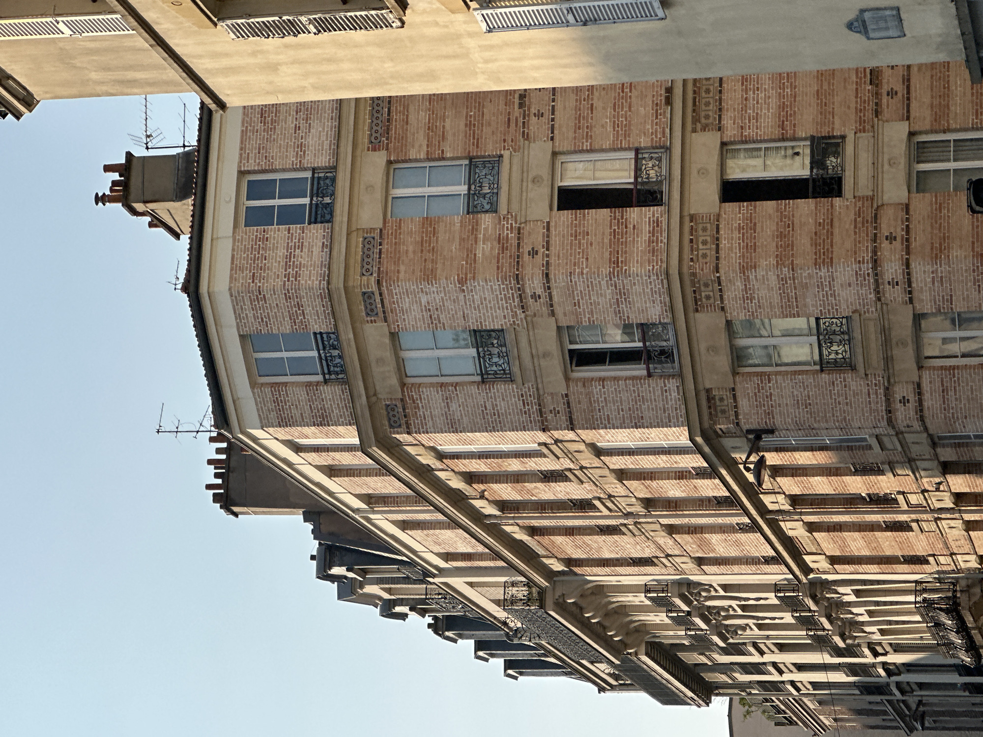 This image shows a view of a tall, multi-story brick building in Paris, France, with a lighter-colored building to its right. The building in the foreground is on a corner, with its facade curving to the left. It has many windows, some with balconies and decorative ironwork. Chimneys and antennas are visible on the roof. The architecture suggests a Haussmannian style, common in Paris. The sky is clear and blue. There are no people visible in the image. The lighting indicates it is daytime, possibly late afternoon or early morning due to the shadows.