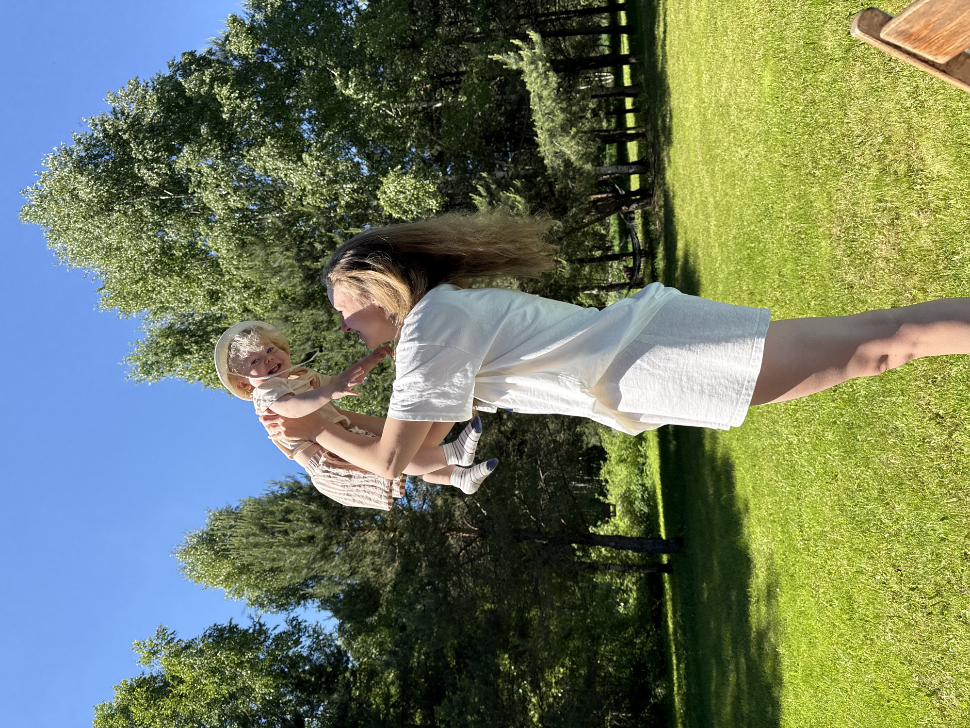 The image captures an outdoor scene on a clear, sunny day in Копнино, Russia. An adult female is positioned right of center, facing left and upward, holding an infant/toddler aloft. The adult female has long, light brown hair and is dressed in a white short-sleeved top and white shorts. The infant/toddler, with light, curly hair and a light-colored bucket hat, wears a light-colored top, striped shorts, and striped socks. The child faces right and downward, interacting with the adult. Both individuals have upward-turned mouths, indicative of smiles. The ground is covered with lush green grass, illuminated by sunlight, with visible shadows. A dense line of green trees, including both deciduous and evergreen species, forms the background beneath a clear blue sky. A hammock is faintly discernible among the trees in the middle distance. A wooden object, possibly a chair arm, is partially visible in the bottom right corner.
