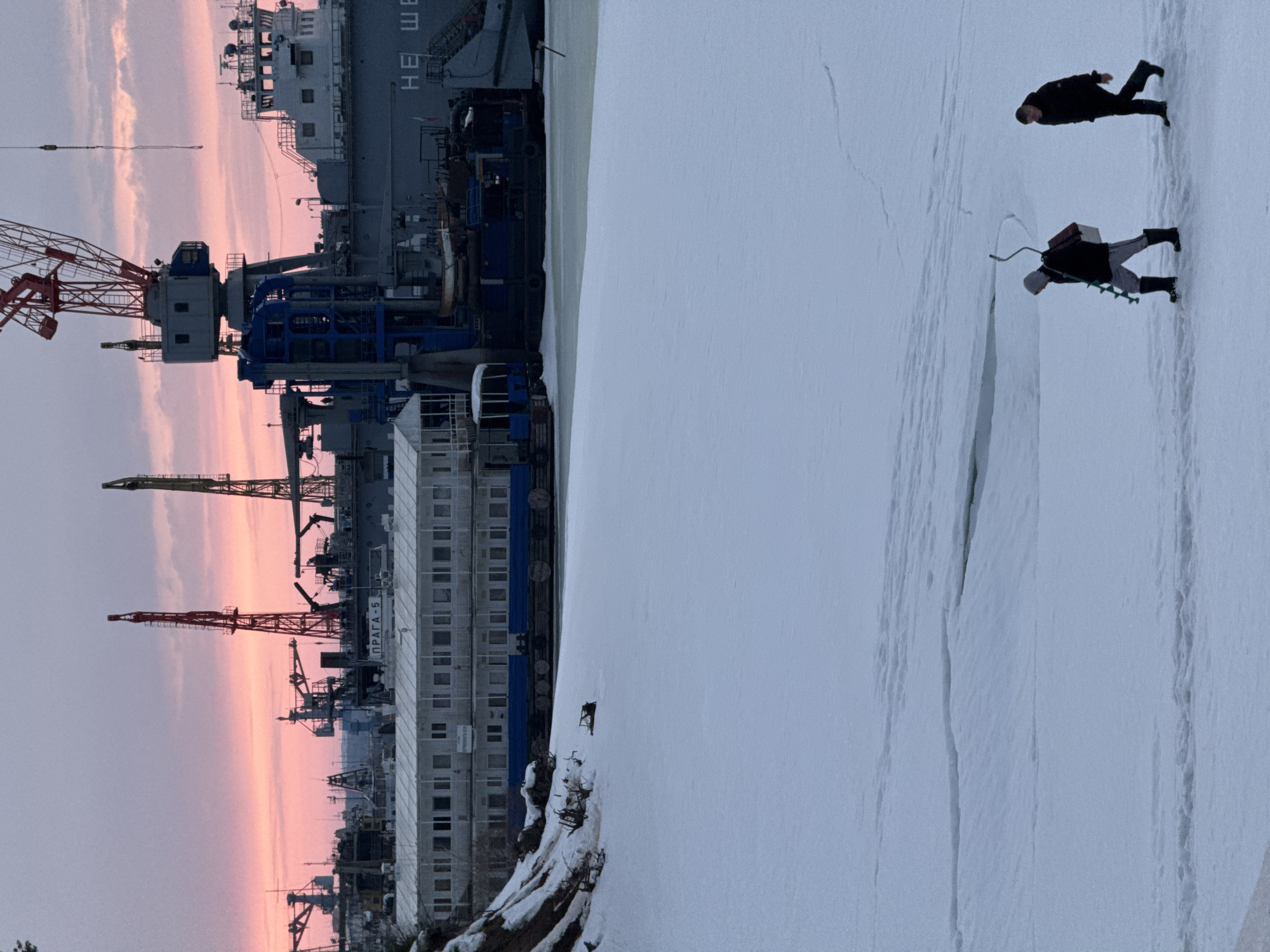 The image depicts a winter scene in Chaikovsky, Russia. In the foreground, two figures are present on a snow-covered, potentially frozen, body of water. One figure is equipped with what appears to be an ice drill and a fishing box. The second figure is walking ahead. Cracks are visible on the snowy surface.
In the background, industrial infrastructure is visible including multiple cranes, buildings, and a structure with "ПРАГА-5" inscribed on it. A large vessel or dockyard is also present. The sky shows a gradient of colors suggesting either a sunrise or sunset.