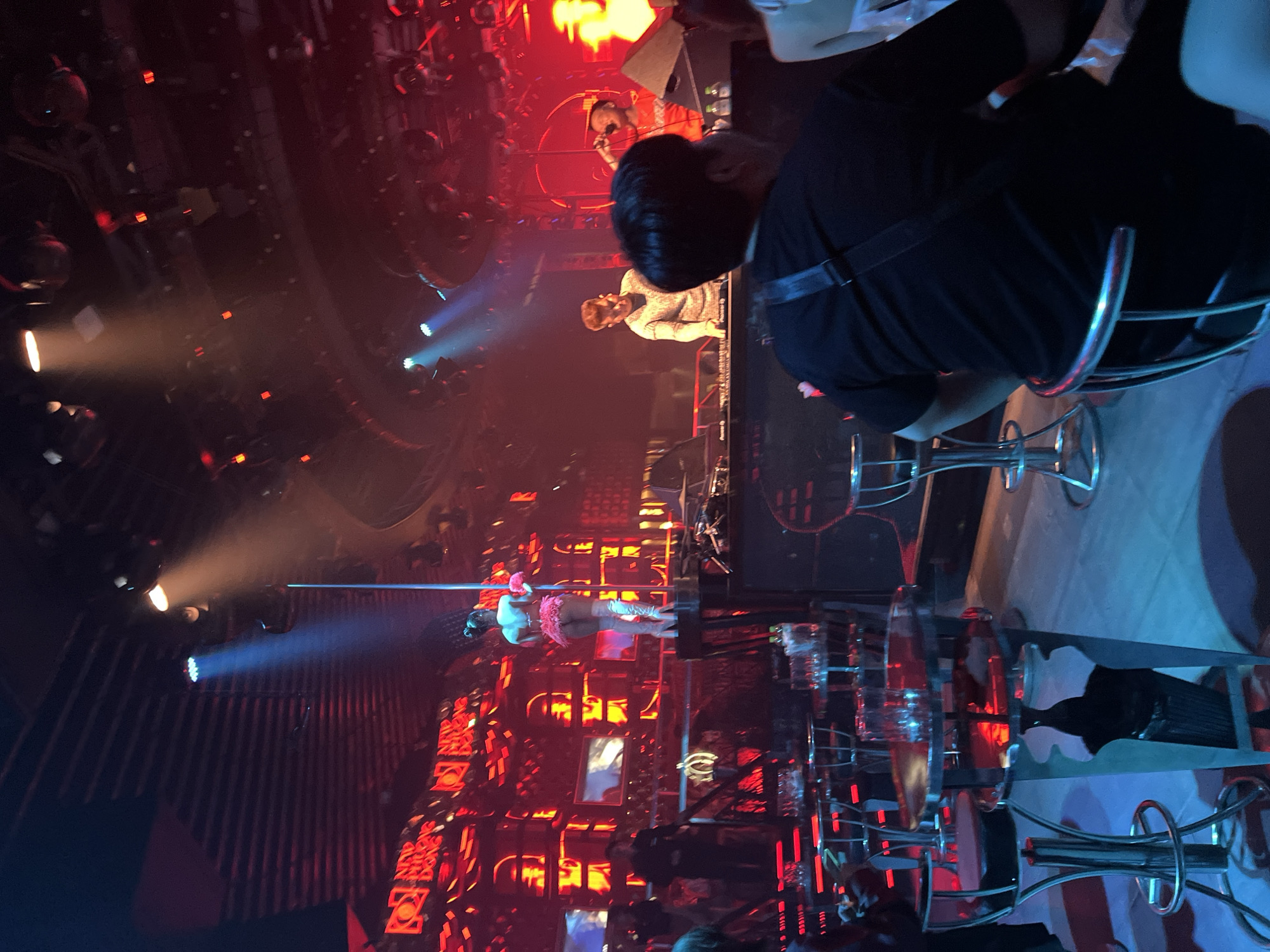 This image depicts the interior of a nightclub in Đà Nẵng, Vietnam. In the foreground, a person wearing a black t-shirt sits facing away from the camera, with their back to the viewer. The club features a stage with a pole dancer in a pink outfit, wearing white boots. Behind the dancer, the walls are adorned with red, illuminated signage. To the right of the stage, DJs are visible at a console, with one man wearing a patterned shirt and another in an orange shirt. Several high-top tables with stools are scattered throughout the venue, some with glasses on them. The lighting is predominantly red and blue, with spotlights illuminating the stage. The overall atmosphere suggests a lively entertainment venue.