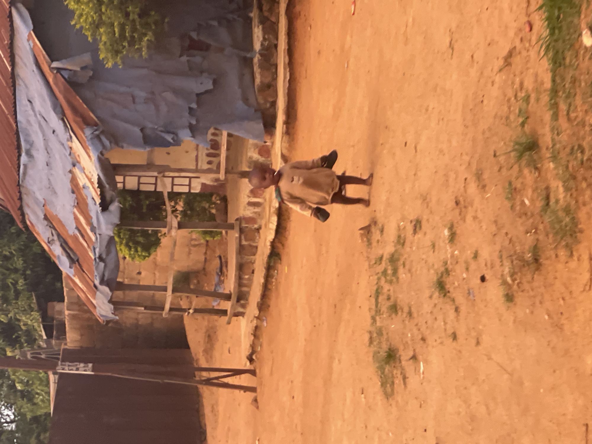 A young child walks down a dirt road in Barikin Gidar Kanat, Nigeria. The child is barefoot and dressed in light-colored clothing, holding what appear to be shoes in their hands. To the left, a building with a partially collapsed roof is visible, along with simple wooden supports. The environment is dry and dusty, with scattered vegetation on the right side of the road.