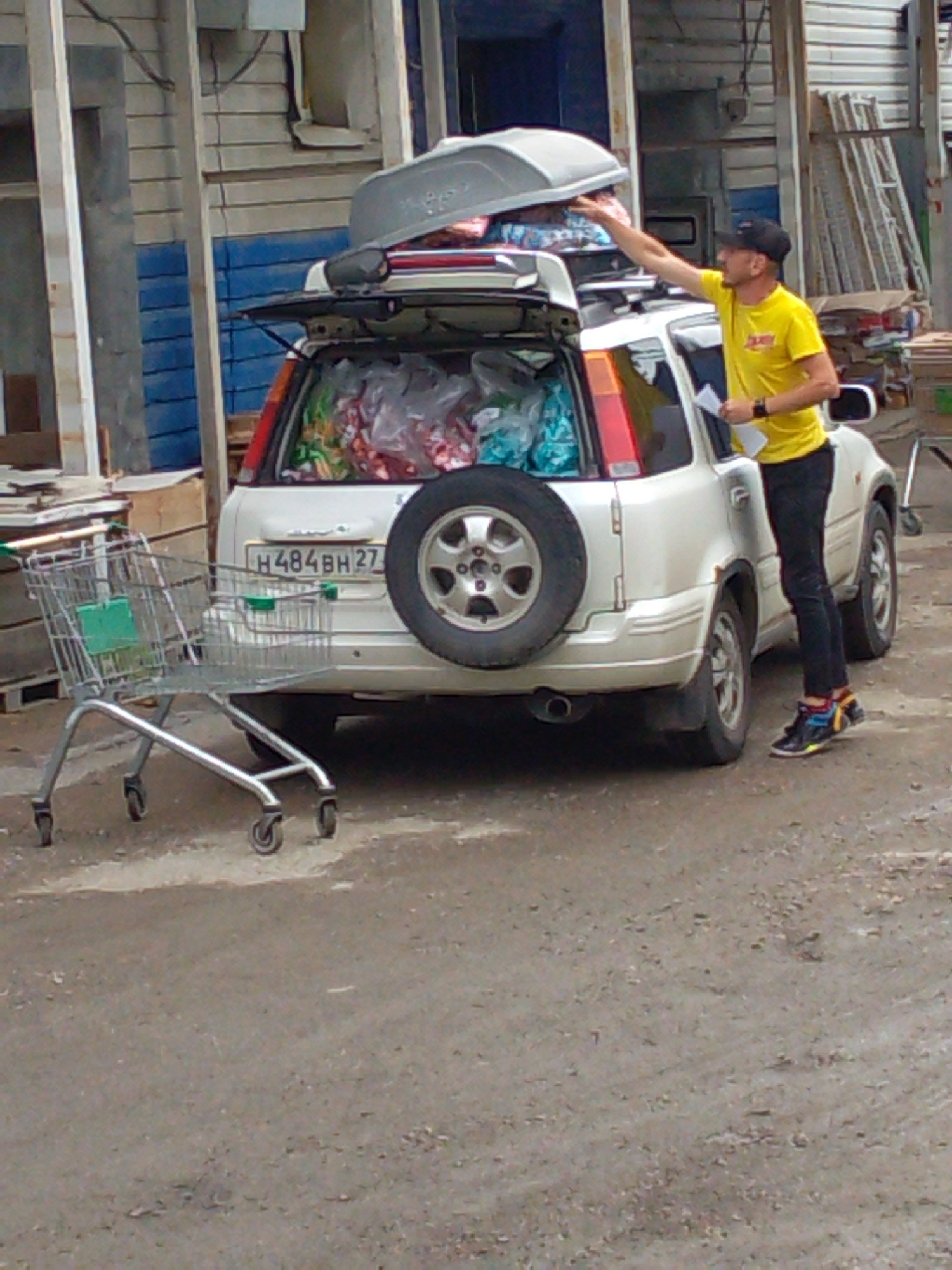 A man wearing a yellow t-shirt, black pants, and a black baseball cap is standing next to a white SUV. The SUV's rear hatch and roof cargo carrier are open. The man is reaching towards the cargo carrier. The vehicle has a license plate that reads "H484BH 27". A shopping cart is positioned to the left of the SUV. The scene appears to be outdoors in an urban setting, possibly a parking lot or street, with buildings and stacked materials in the background. The ground is dirt and gravel. The overall impression is of someone loading or unloading the vehicle.