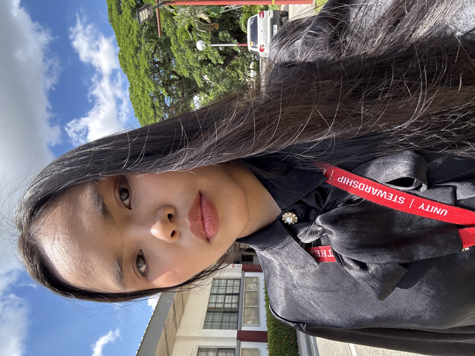 A close-up, eye-level outdoor shot looking up at a young Southeast Asian woman with long, dark hair. She is wearing a black silky shirt with a decorative pearl button and a red lanyard imprinted with the words "UNITY | STEWARDSHIP". The background consists of a building, foliage, a white vehicle, and a blue sky with clouds. This picture was taken at Ateneo de Davao University in Davao City, Philippines.
