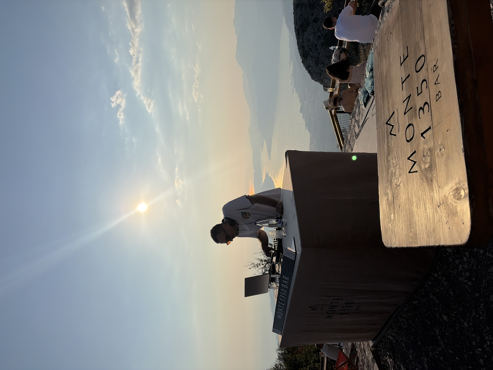 The image captures a DJ set at Monte 1350 Bar, a mountaintop establishment in Kuk, Montenegro, during sunset. A man, presumably the DJ, stands behind a counter with a laptop and DJ equipment, wearing sunglasses and a white t-shirt. Several patrons are visible in the background, seated at tables and enjoying the view of the bay and surrounding mountains. The bar counter itself is branded with "MONTE 1350 BAR." The scene is illuminated by the warm glow of the setting sun, casting long shadows and creating a serene atmosphere.