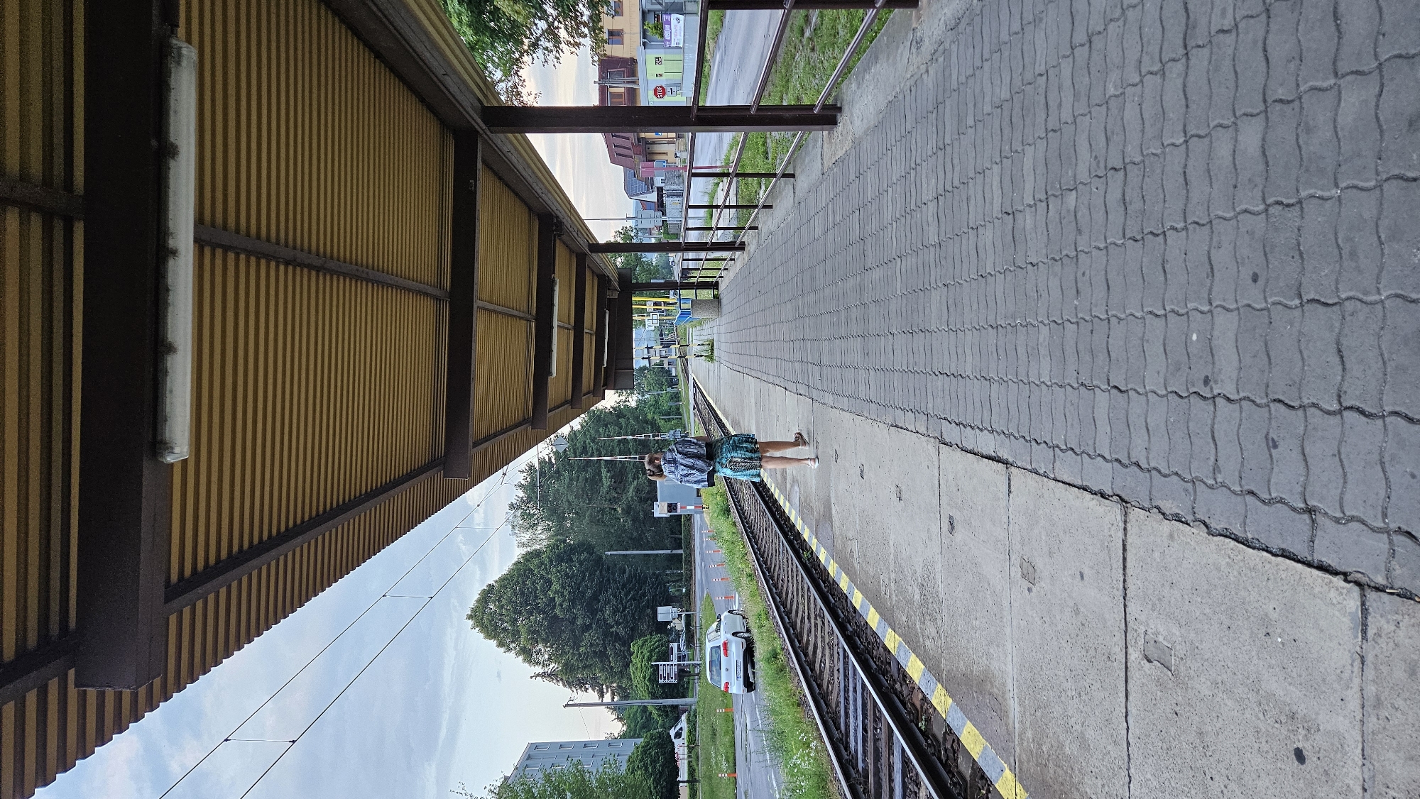A full-shot, vertical, outdoor, daylight image captures a person walking away from the camera down a train station platform. The person is wearing a backpack and a patterned skirt and appears to be walking towards a distant train track. The platform is paved with interlocking gray stones and has a concrete edge next to the train tracks. The train tracks are on the left side of the image, with a white car and other vehicles visible on a road beyond the tracks. The station has a yellow corrugated metal roof supported by dark brown beams, and a fluorescent light fixture is visible attached to the underside of the roof. Buildings and trees line the background. The image is set in the city of Prešov, Slovakia, at a tram stop.