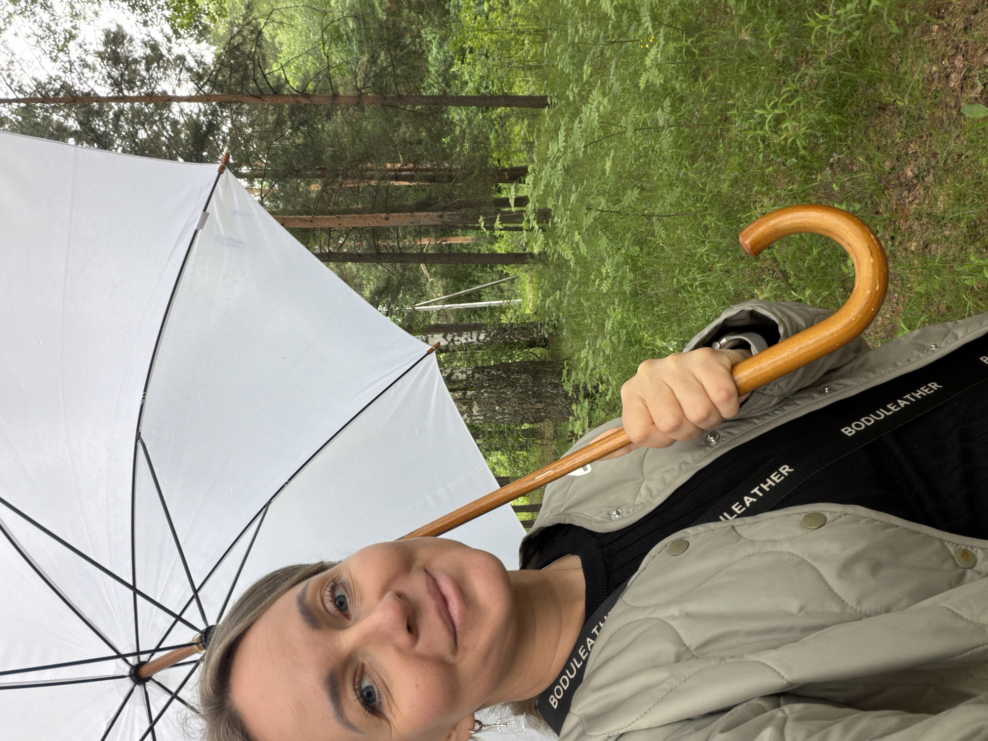 A selfie-style portrait captures a woman holding a white umbrella in a forest setting in Chaikovsky, Russia. The image is taken at a slight upward angle and is rotated 90 degrees counter-clockwise.
The woman is in the foreground, looking directly at the camera with a subtle smile. She has light-colored hair and blue eyes. She is wearing a light gray, possibly quilted, jacket over a dark, ribbed top. A black strap with the white text "BODULEATHER" is visible across her chest. Her right hand holds the curved, wooden handle of the open white umbrella, which extends across the left side of the frame. The umbrella's structure, with its black metal ribs, is clearly visible against the white canopy.
Behind the woman and the umbrella, a dense forest fills the background. Tall trees with visible trunks and abundant green foliage create a lush, natural environment. The ground is covered with rich green undergrowth. The presence of the umbrella suggests potentially overcast weather or a recent rain, though no active precipitation is visible. The scene evokes a calm, outdoor moment in nature.