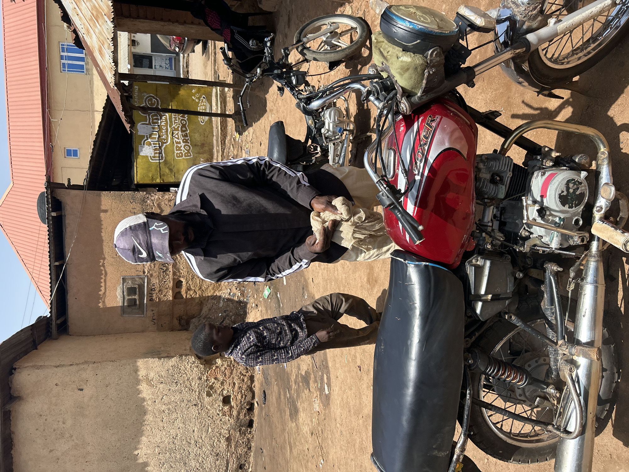 In Jos, Nigeria, a young boy stands beside a red motorcycle as a man, wearing a black long-sleeved shirt with white stripes and a black baseball cap with a white Nike logo, cleans his hands with a light-colored cloth. The scene unfolds on a dirt ground with buildings and a corrugated metal roof in the background. A yellow sign with text that appears to say "CHINCHIN BREAK THE BOREDOM" is visible. The red motorcycle features the word "BOXER" on its fuel tank. Another motorcycle is partially visible behind the man. The overall environment suggests an outdoor marketplace or a residential area with a rough, unpaved surface.