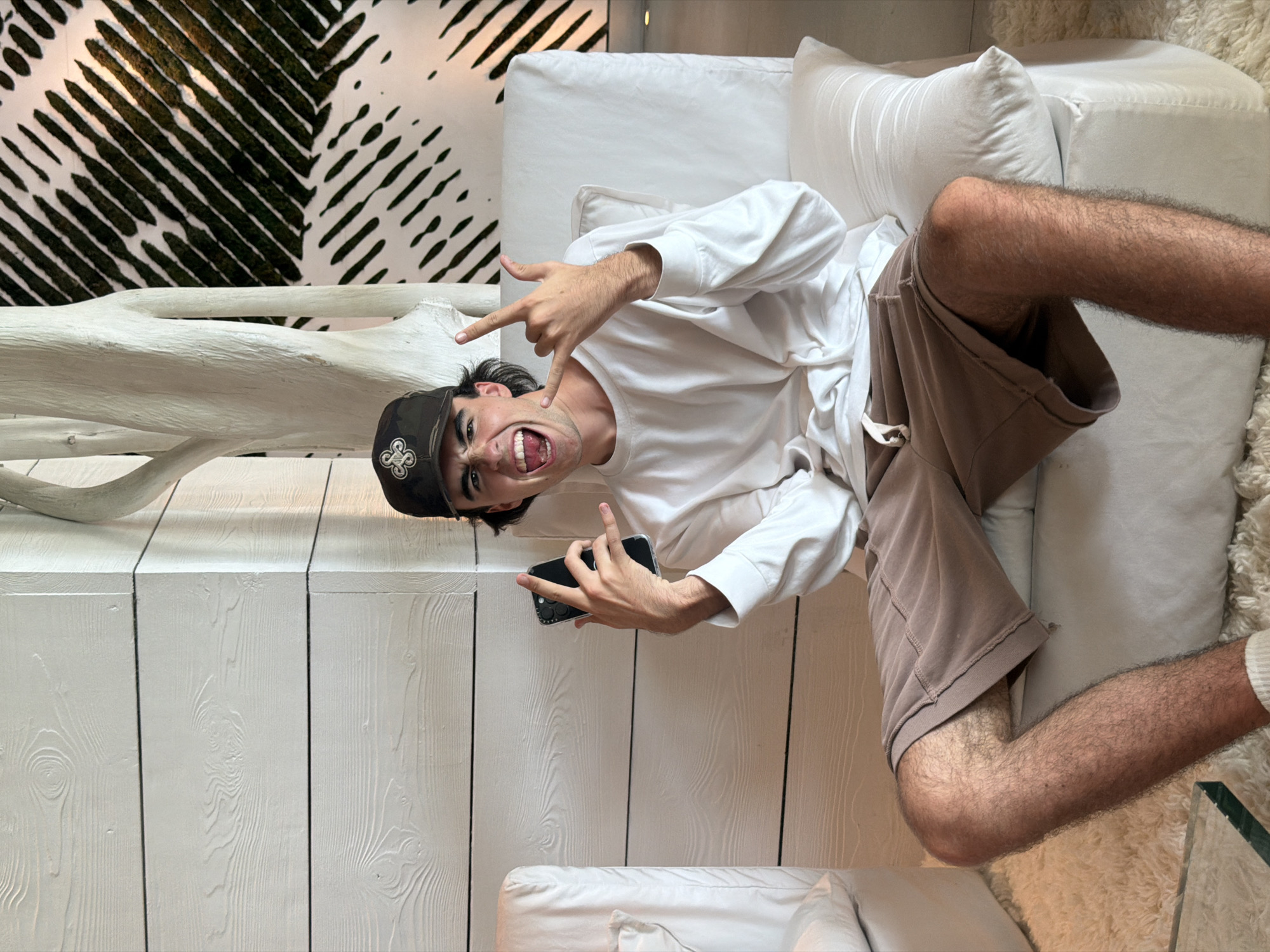 A young man is captured in a dynamic, vertically oriented indoor shot, appearing to lie on a white sofa. He looks directly at the camera with an open-mouthed, excited expression, holding both hands up in a "rock on" or "devil horns" gesture. He wears a dark baseball cap with a distinct white logo, a plain white long-sleeved t-shirt, and light brown shorts, revealing noticeably hairy arms and legs. In his left hand, he holds a black smartphone, showcasing its multi-lens camera system.
The background features a clean, modern interior. To the man's left, a large, organic-shaped white sculpture, resembling bleached wood or coral, stands prominently. Behind him, a wall is paneled with white textured wood. Above this, a striking black and white abstract pattern of diagonal lines and shapes covers another wall section. Parts of a fluffy white rug and a glass table edge are visible near his legs. The setting suggests a bright, contemporary space, possibly in Miami Beach, United States of America. The man's pose and expression convey a playful and energetic mood.