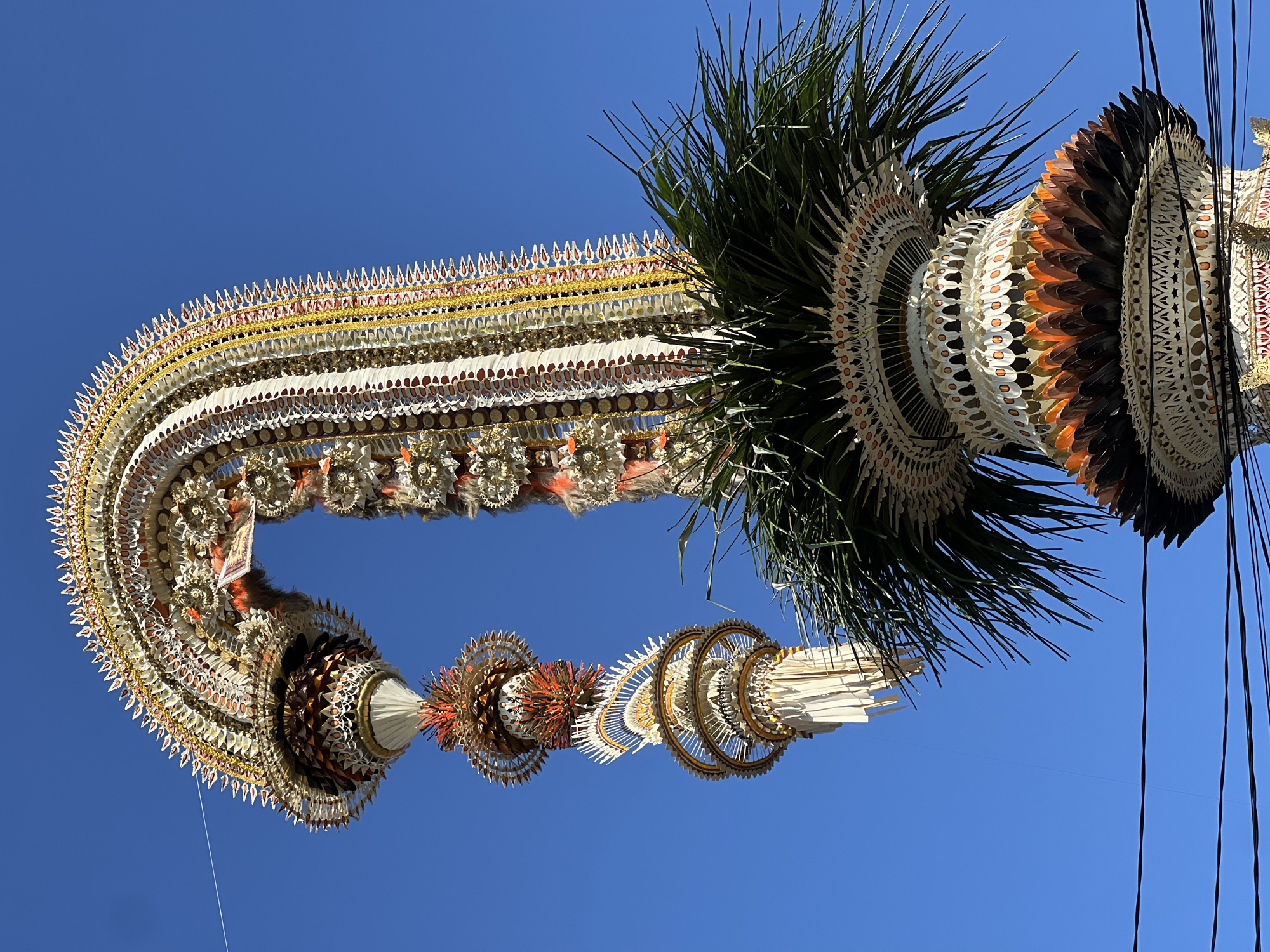This media file features an elaborate, festive decoration, likely a "penjor" or similar ceremonial structure, against a clear, bright blue sky. The decoration is suspended and appears to be made of natural materials like palm fronds and possibly feathers or carved wood, meticulously arranged in intricate patterns. The structure has a complex, layered design with circular motifs, a prominent green frond section, and accents of white, gold, and reddish-brown. It curves dramatically, resembling a horseshoe or a large, ornate garland. Thin wires or strings are visible, suggesting it is hung or supported, and power lines are also present in the background. The bright sunlight indicates it is daytime, likely mid-morning or afternoon given the clear sky. The overall impression is one of celebration and cultural artistry, commonly seen during religious or cultural events in Indonesia, specifically in regions like North Kuta. There is no visible text.