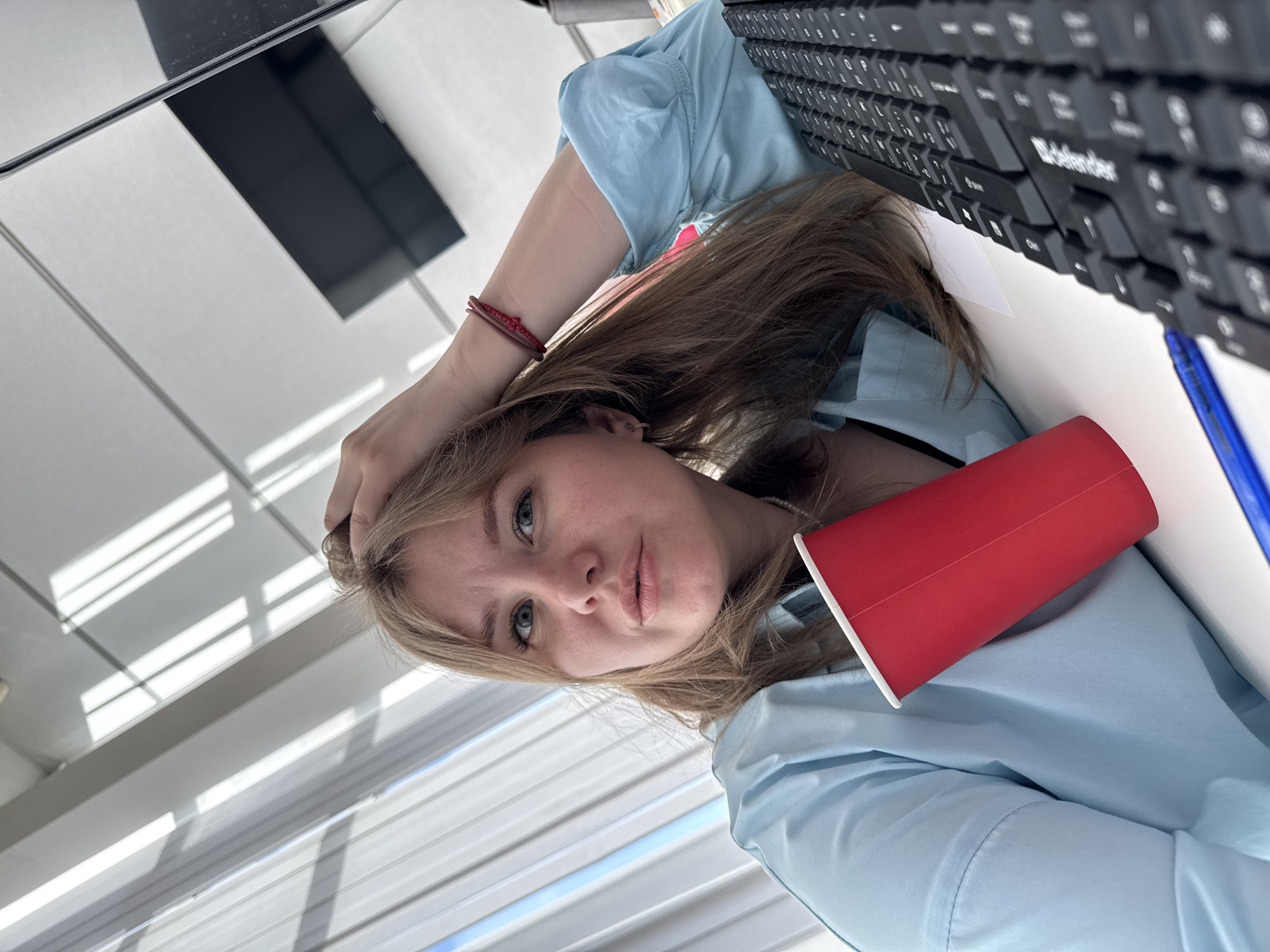 A young, light-skinned woman with blonde hair sits at a desk. She wears a light blue collared shirt. Her left hand rests on her head, and her facial expression is neutral. A red paper cup and a black keyboard are on the desk. A blue pen lies to the right of the cup. The scene is indoors, near a window with horizontal blinds. According to metadata, the location is Perm, Russia.