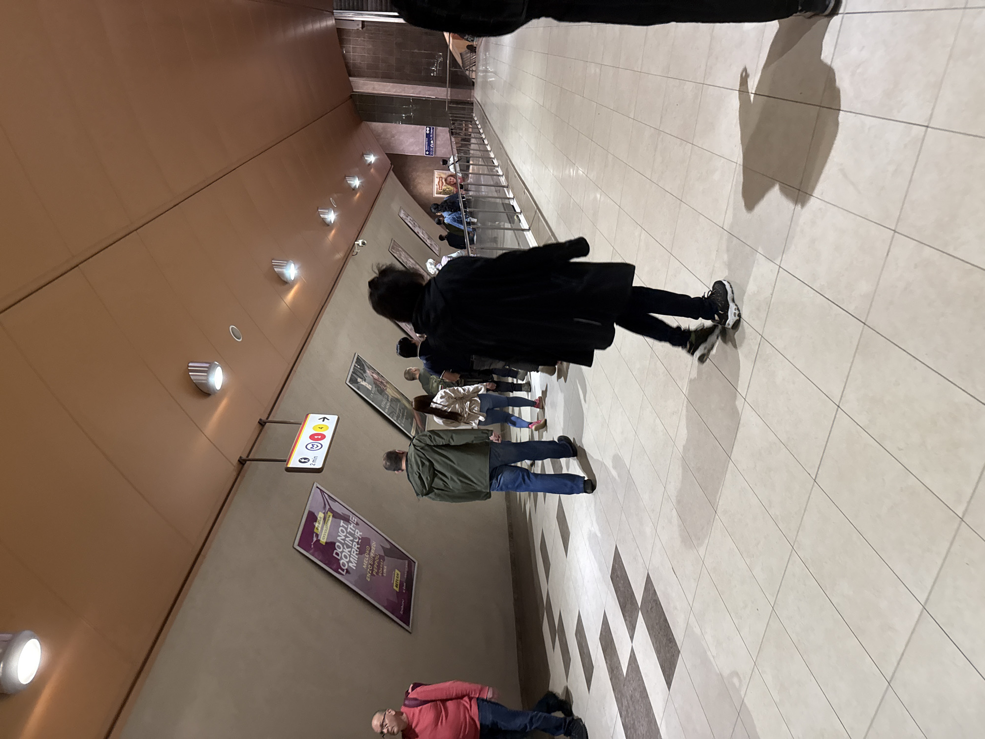 A photograph depicts an indoor setting in Sofia, Bulgaria, showcasing a subway passage area. The scene is populated with several people walking in the same direction. The foreground features a person wearing a black coat and dark trousers, walking away from the camera. Adjacent to them, slightly further into the scene, a man in a green jacket and blue jeans is also progressing forward. Beyond these individuals, several more people can be observed, seemingly grouped together.
The environment includes subway-related signage mounted on the walls, indicating directions and travel times. Specifically, a sign reads "2 min," possibly indicating a walking time to a specific platform or destination. Advertisements are displayed on the wall, including one with the phrase "DO NOT LOOK IN THE MIRROR," featuring a list of names beneath it. The architecture consists of brown walls with recessed lighting fixtures and a tiled floor.