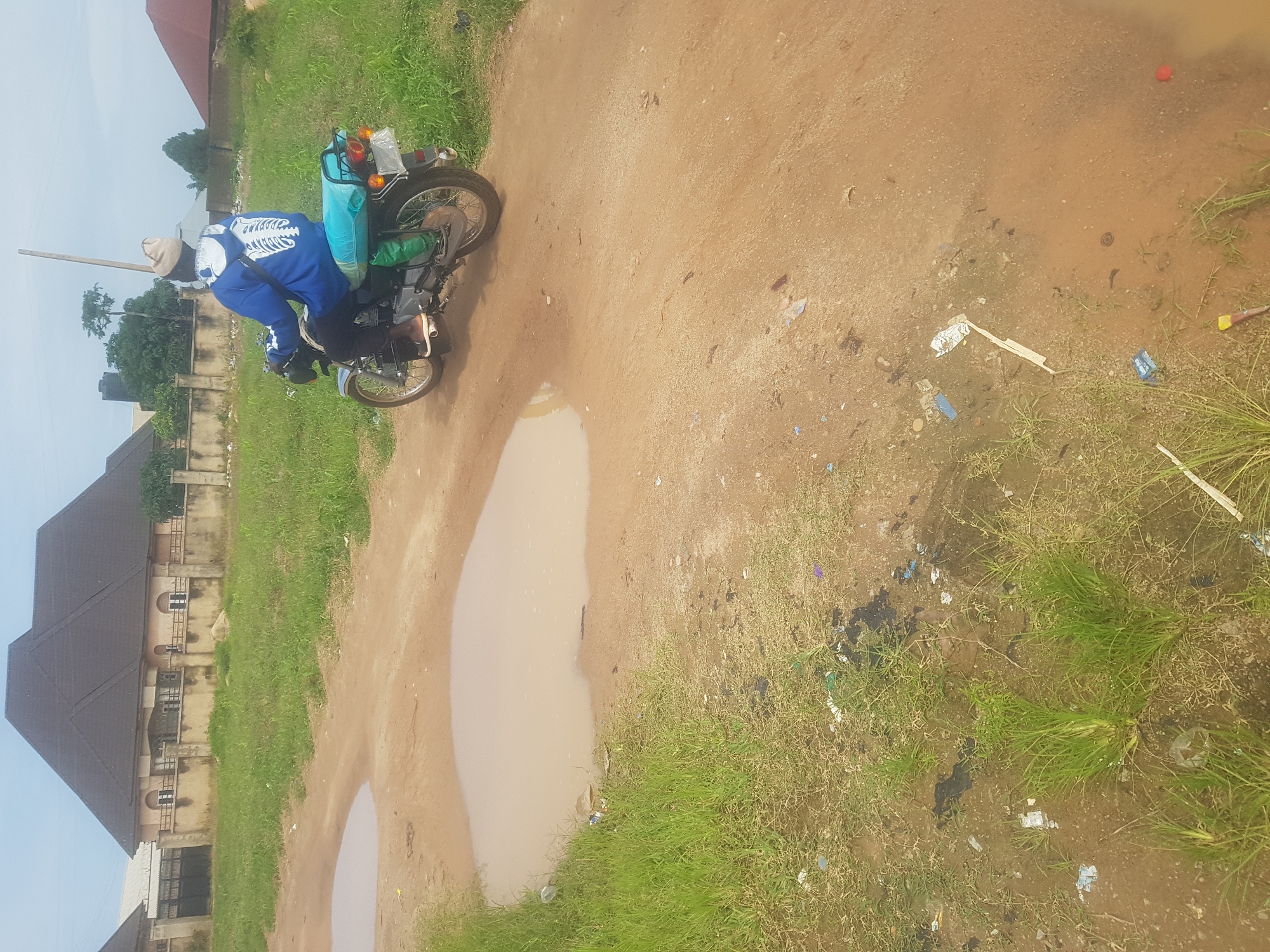 A person wearing a blue hoodie with a white skeleton design on the back and a beige beanie rides a silver motorcycle. The motorcycle has a turquoise seat cover. The rider is on a dirt road with several large puddles. In the background, there is a fence and a large house with a dark roof. The environment is overgrown with grass and appears to be a rural or suburban area. There is scattered litter on the ground. The sky is overcast.