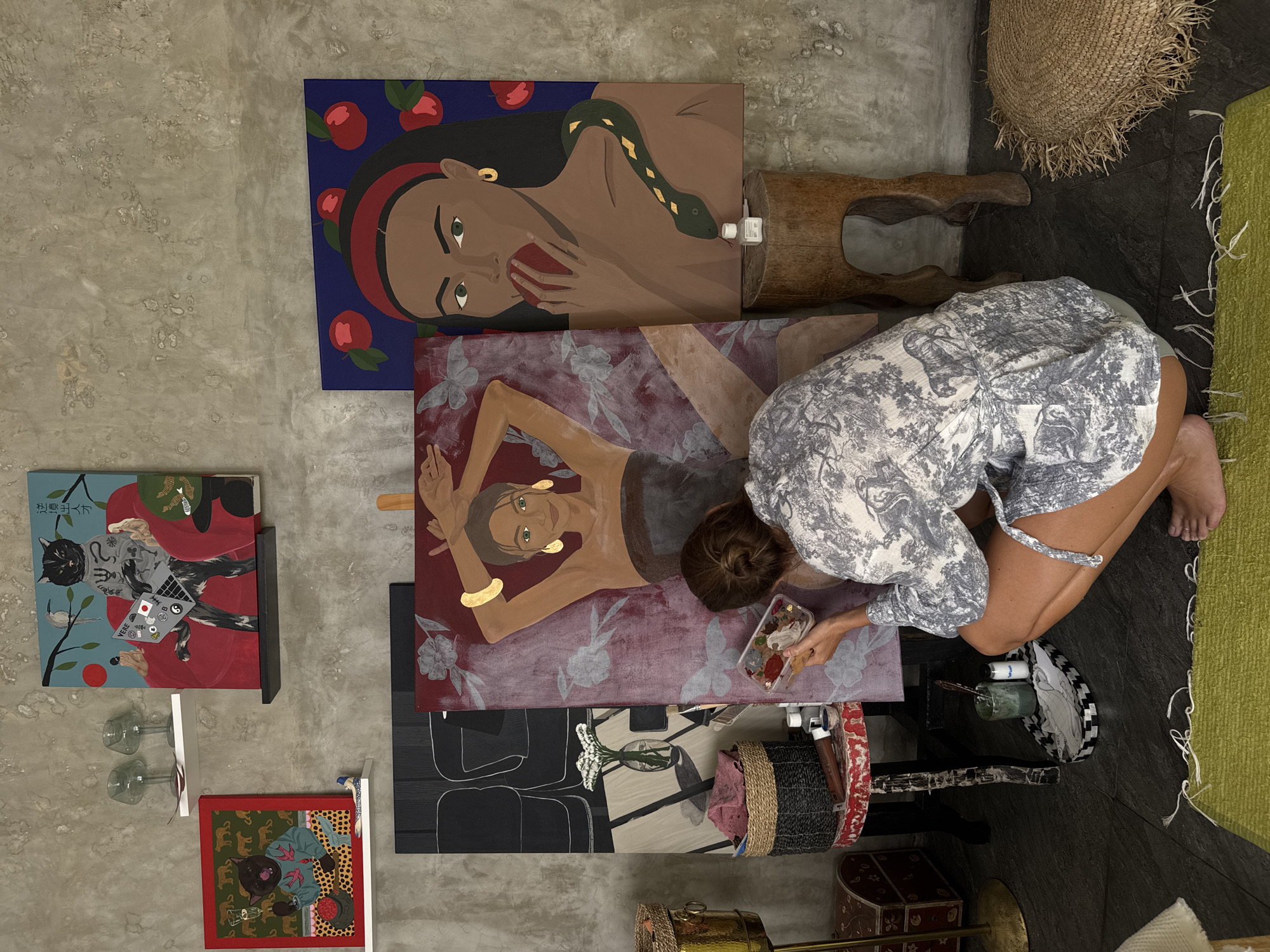 An overhead shot captures a woman painting on the floor, surrounded by vibrant artworks and art supplies, within what appears to be an indoor studio or creative space. The floor is a mix of light grey concrete and darker grey tiles, with a lime green shaggy rug partially visible on the right.
The central figure, a woman with brown hair tied in a bun, is kneeling or crouching, focused on her work. She wears a light-colored top with an intricate pattern and shorts, her bare feet visible. In her hands, she holds a transparent paint palette and a brush, actively applying paint to a canvas positioned in front of her.
Several paintings are scattered around. Directly in front of the woman is a medium-sized canvas depicting a reclining woman with dark skin, green eyes, gold hoop earrings, and a gold arm cuff, against a reddish-purple background adorned with subtle floral and butterfly patterns. Above this, a larger painting shows a dark-skinned woman with a red headband, green eyes, and a green snake with golden markings draped around her, holding a red apple to her mouth. This artwork features a dark blue background with red apples.
To the left, smaller framed paintings are visible. One depicts a black cat wearing clothes and holding a laptop, with Japanese text reading "松尾芭蕉" (Matsuo Bashō) and "YERE". Another portrays a clothed brown cat holding a drink and a strawberry against a red and yellow polka-dotted background.
Various objects are interspersed throughout the scene: a rustic wooden stool, a large fringed straw basket, a smaller patterned basket, a white bottle (possibly hand sanitizer) on the stool, a glass with paintbrushes, two empty wine glasses, a decorative red wooden box, and a brass object. The scene suggests a dedicated artistic activity in a casual, bohemian setting.