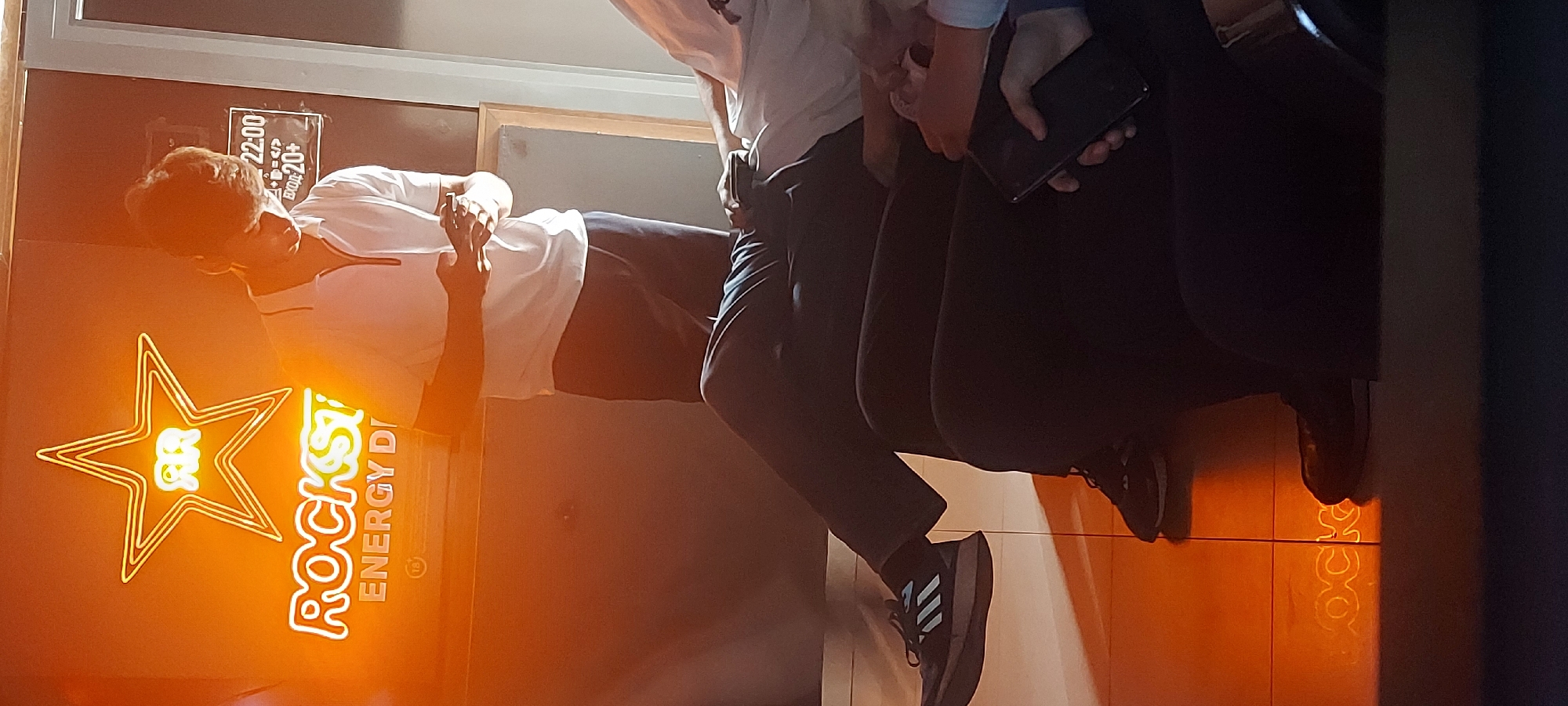 Here's a description of the image:
**Scene**: The image appears to be an indoor shot in what looks like a bar or club in Tashkent, Uzbekistan.
**People**: Two people are visible. The first person is standing in front of a wall that has a Rockstar Energy Drink sign. They're wearing a white shirt and dark pants. The other person is partially visible, wearing dark clothing, and is holding a smartphone. There's a sticker on the wall that looks like it says "22:00" and "+20".
**Objects**: A bright neon Rockstar Energy Drink sign stands out. The sign is on the wall and it is a yellow orange color. Additionally, there is a person holding a phone.
**Actions/Interactions**: The person in the white shirt seems to be interacting with the other person, who is holding the smartphone.