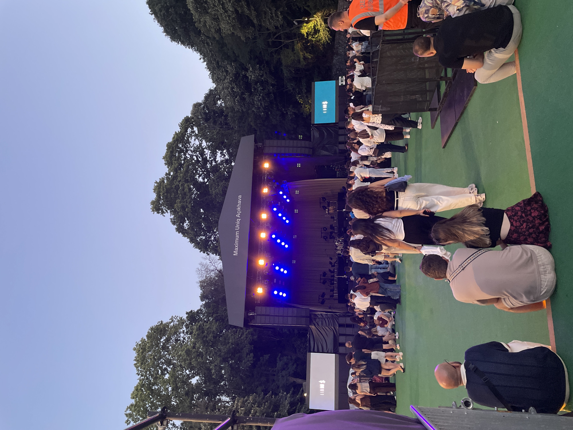 The image depicts an outdoor concert venue in Sarıyer, Turkey. A stage is set up with bright blue and orange lights illuminating a dark backdrop. The venue is filled with a large crowd of people, many standing and watching the stage, while others are seated on the green ground. In the foreground, several individuals are seated, facing away from the camera towards the performance area. Among the seated figures, there is a bald man wearing glasses and a dark blue shirt, and a man in a light brown shirt. To the right of the stage, a large screen displays a graphic with white lines and symbols. A security guard in an orange vest is visible on the far right of the image. The background is dominated by lush green trees, suggesting the concert is taking place in a natural setting within the city. The sky is a clear, pale blue, indicating it is either late afternoon or early evening.