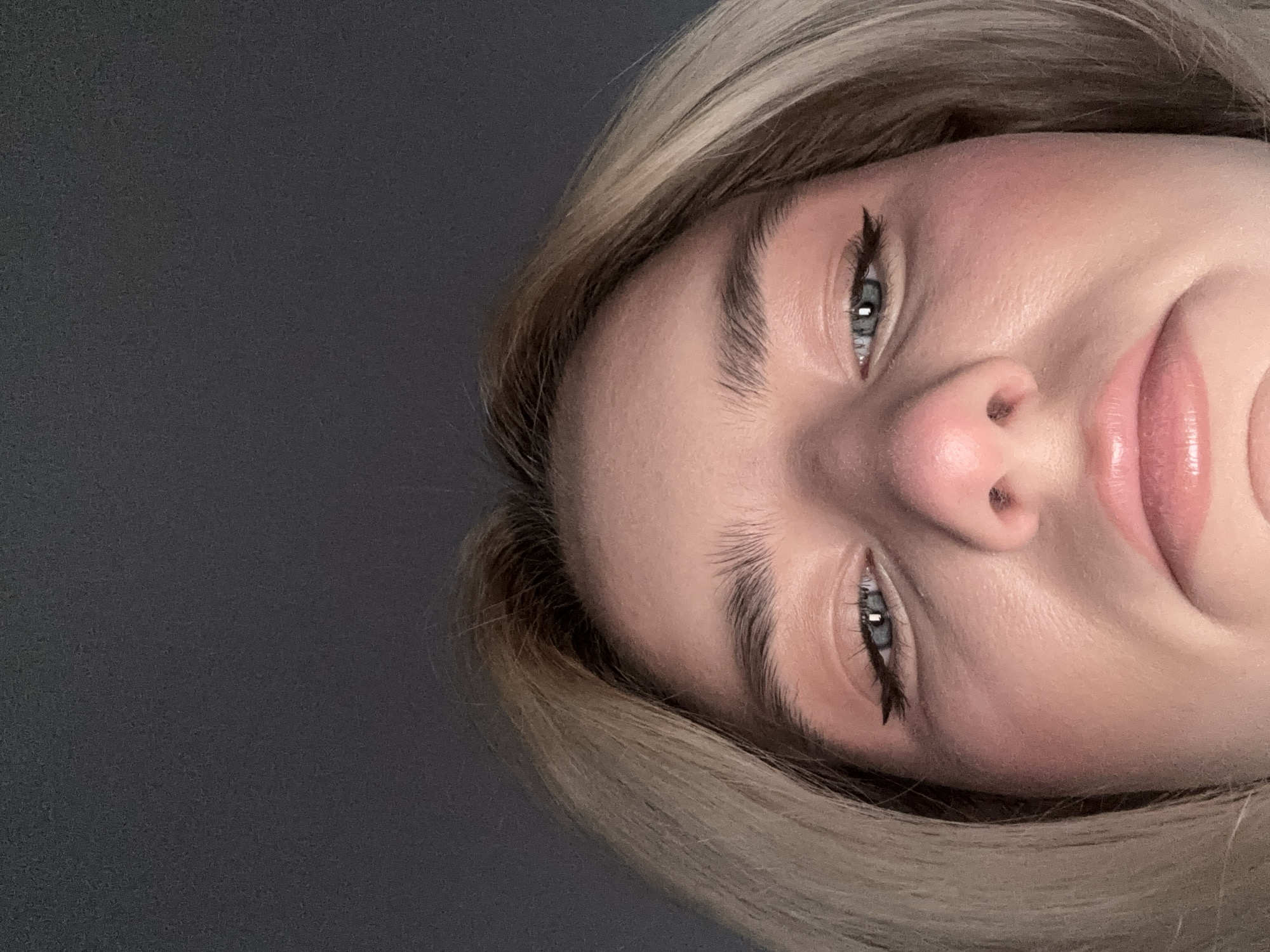A close-up, angled shot of a woman's face. The woman has fair skin, blonde hair, and light blue eyes. She has eyeliner applied and her lips appear to have lip gloss on. The background is a plain, dark gray. The image does not show any indication of being in Vsevolozhsk, Russia or any other specific location.