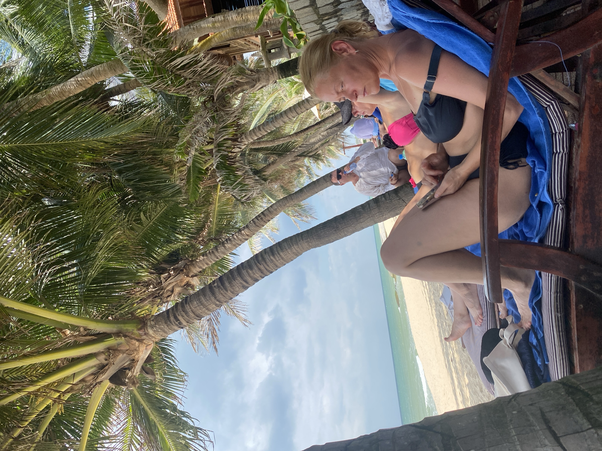 In Phu Thuy Ward, Vietnam, on a sunny day at the beach, two individuals are relaxing on lounge chairs under the shade of palm trees. A woman in the foreground is wearing a black bikini and holding a phone, while a man in the background is drinking from a bottle. The ocean and sandy beach are visible behind them. The scene is captured from a low angle, looking up towards the palm trees and sky.