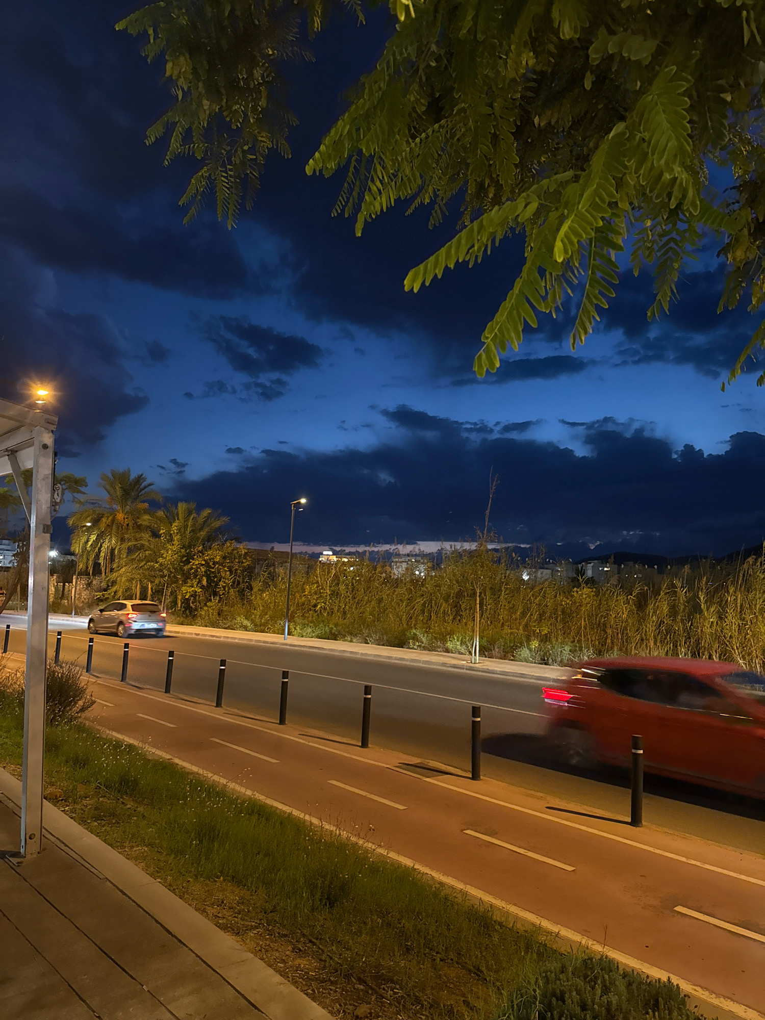 The image depicts a twilight scene on a road in Ibiza, Spain. The sky is a deep blue with dark clouds, suggesting either dusk or dawn. In the foreground, a streetlamp with a warm yellow light illuminates a sidewalk and a grassy area. A road runs parallel to the sidewalk, with a dedicated red bike lane marked by white lines. Black bollards separate the bike lane from the main road. Two cars are visible on the road: a silver car traveling in the left lane and a red car, blurred by motion, in the right lane, indicating it is moving at a high speed. Palm trees and dense vegetation are visible behind the road, with some buildings faintly illuminated in the distance. The overall impression is a peaceful, yet dynamic, urban landscape at the transition between day and night.