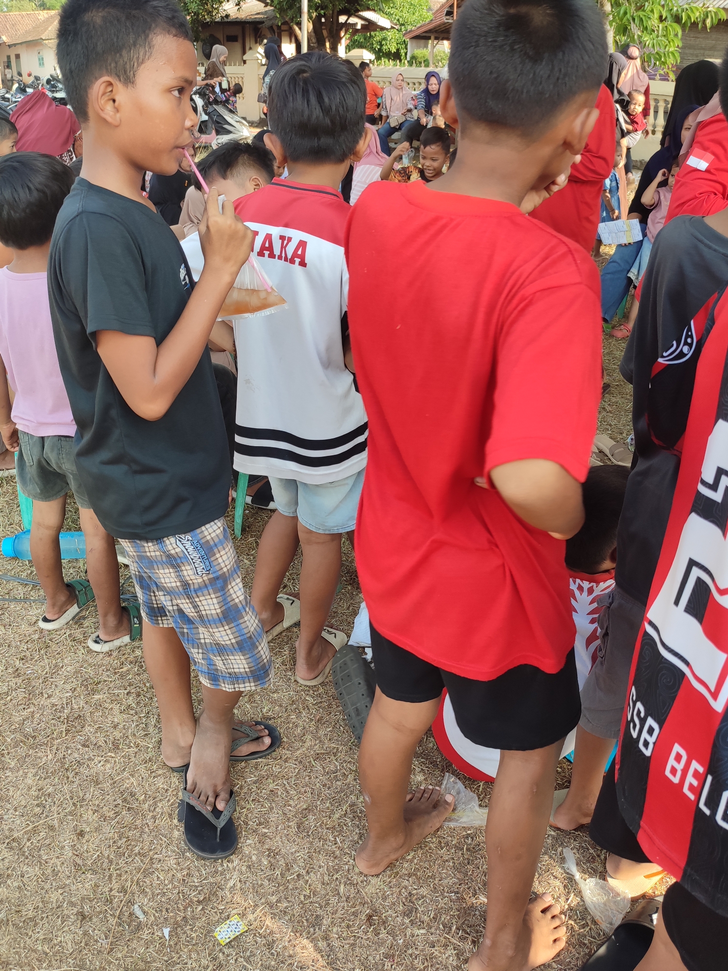 The image depicts a group of children and adults gathered outdoors in what appears to be a community event or gathering. The ground is covered in dry grass and straw. In the foreground, several boys are standing, some wearing t-shirts and shorts. One boy in a black t-shirt and checkered shorts is drinking from a clear plastic bag containing a light-colored liquid, using a pink straw. Another boy to his right wears a red t-shirt and black shorts. Behind them, other children and adults are visible, some seated and some standing. Clothing includes t-shirts, headscarves, and traditional Indonesian attire. Several motorcycles are parked in the background, suggesting transportation. The architecture in the background consists of simple buildings with tiled roofs. The scene is likely set in the city of Nusa Jaya, Indonesia, given the cultural context and presence of motorcycles, which are common modes of transport there. The overall atmosphere suggests a lively, informal gathering.