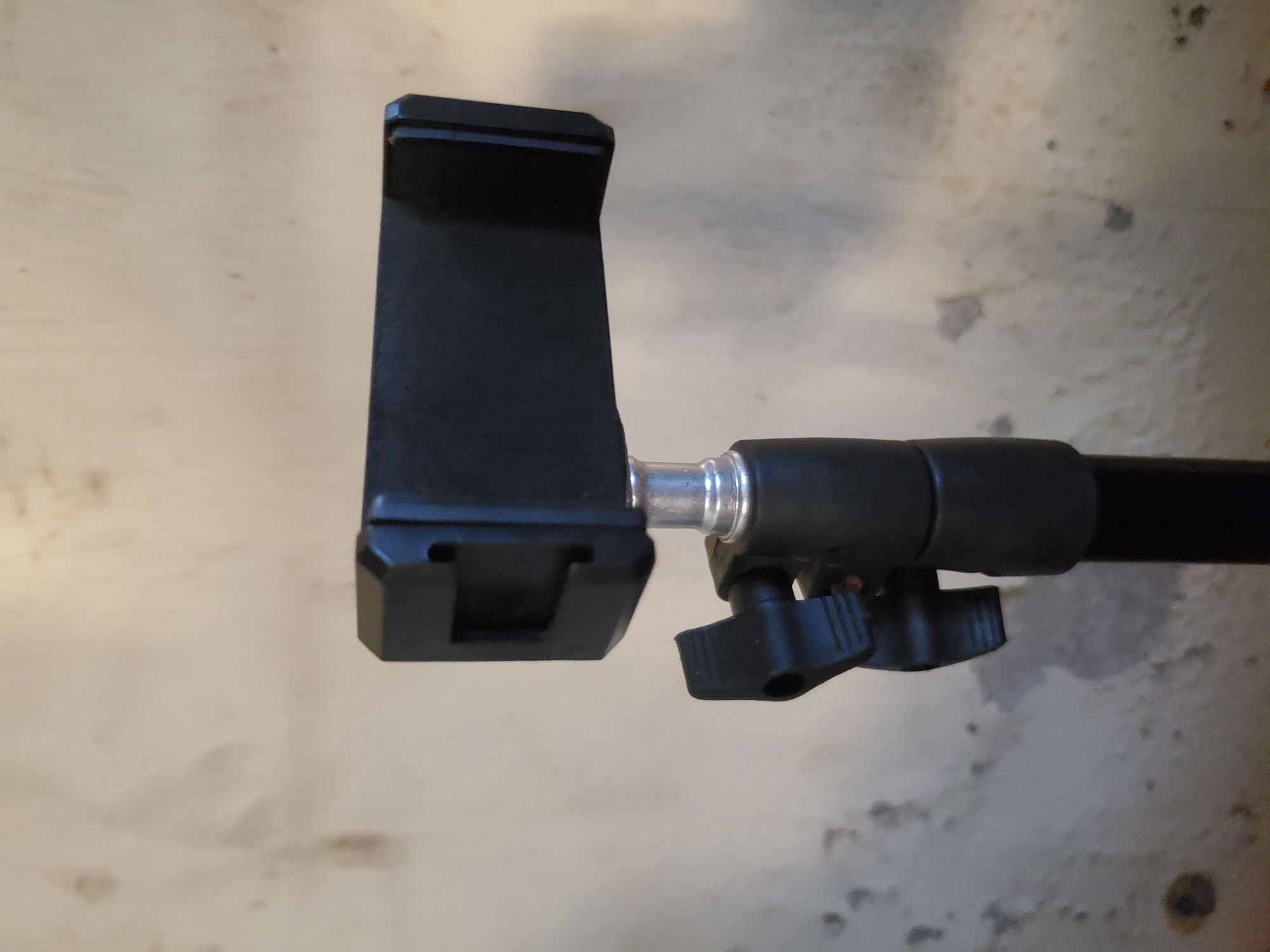 The image displays a close-up of a black smartphone holder or mount attached to a black pole or rod. The holder, which is made of a matte black plastic or metal material, is designed to securely grip a smartphone. It features a spring-loaded or adjustable jaw opening at the top and a fixed lower support.
Connecting the holder to the pole is a silver, cylindrical joint with ridged edges, indicating it might be a threaded or adjustable connection point. Below this joint, on the black pole, are two black, winged adjustment knobs, suggesting that the angle or position of the holder can be altered and secured. The pole extends horizontally out of view to the right side of the frame.
The background is a light-colored, textured surface, possibly a wall or a floor, with visible darker smudges, stains, or mottling, giving it an aged or worn appearance. There are no people visible, and no specific activity is taking place; the equipment is shown in a static state. No discernible text is present on any part of the objects or background. The lighting is neutral, casting soft shadows, but provides no specific cues about the time of day or outdoor weather conditions. The image lacks specific location cues for Mararaba, Nigeria.