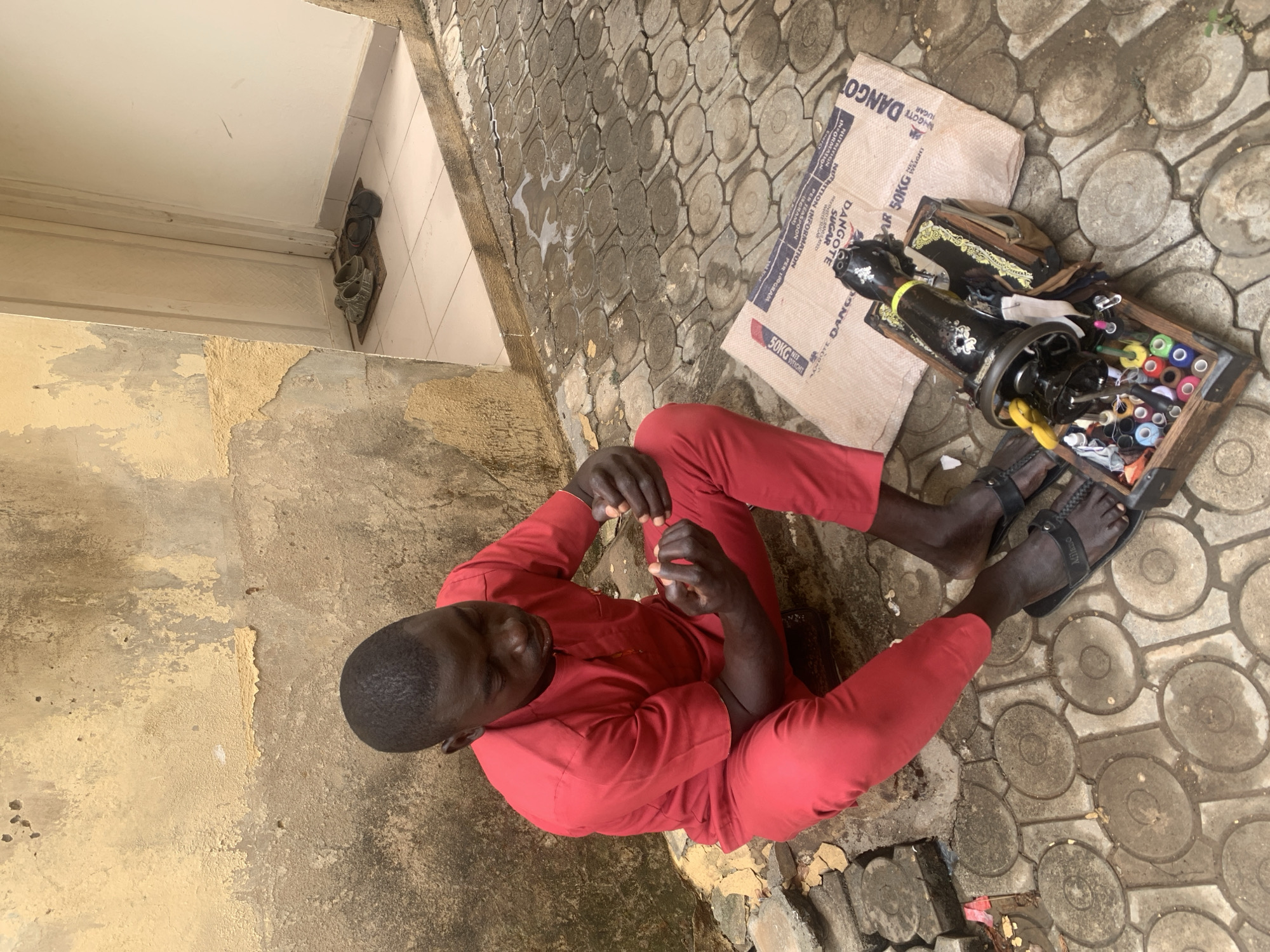 A man in a red outfit sits cross-legged on a patterned brick surface in an outdoor setting. Next to him on a "DANGOTE" brand sugar bag is a black sewing machine with threads and accessories in a wooden tray. The scene includes a building wall with peeling paint and a glimpse of shoes near a doorway. The environment appears to be a simple outdoor workspace. Abuja, Nigeria is not explicitly indicated, however.