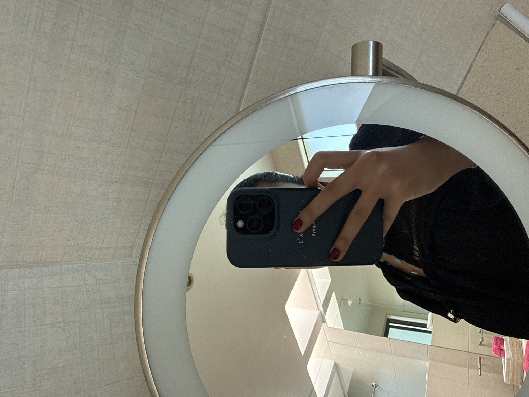 A person holds a dark blue smartphone with three camera lenses in front of a circular, illuminated mirror, reflecting the bathroom setting of a building in San Bernardino Tlaxcalancingo, Mexico. The person has manicured nails with dark red polish. A black, lacy garment covers part of their torso. The bathroom features beige tiles, a window, and pink towels.
