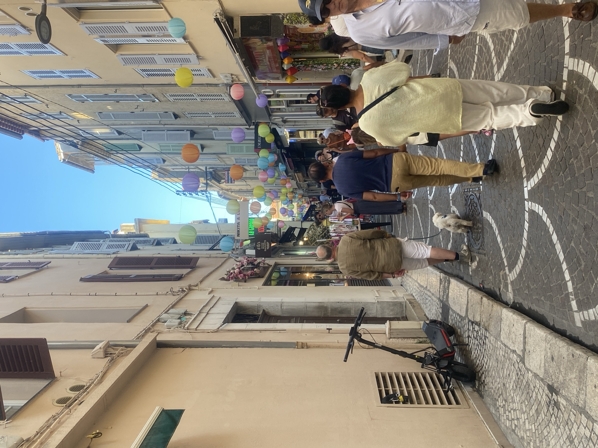 The image depicts a street scene in Antibes, France, likely in the old town, given the architecture and narrow pedestrian alley. The street is paved with cobblestones and lined with buildings featuring traditional shutters and stucco facades. Paper lanterns in various colors are strung across the street, creating a festive atmosphere.
Several people are walking along the street. In the foreground, a man with a shaved head wearing a brown short-sleeved shirt and white shorts walks away from the camera, holding a leash attached to a small white dog. To his right, a woman in a pale yellow top and white pants walks away from the camera, with a black strap across her shoulder. Further to the right, another person in a light blue shirt and white skirt also walks away from the camera. In the background, more individuals are seen strolling, some appearing to be browsing shops.
On the left side of the street, an electric scooter is parked against a building wall next to a ventilation grate. The buildings house shops with displays visible through their windows, one of which appears to be a beauty salon. The overall scene suggests a lively and popular shopping and walking area during daylight hours under a clear blue sky.