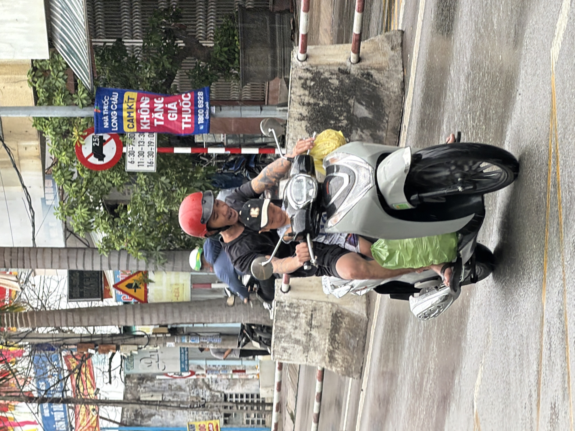 A man and a young boy are riding on a silver scooter on a wet, paved road in Nha Trang, Vietnam. The man, wearing a red helmet and a dark t-shirt, is driving the scooter. The boy, wearing a black baseball cap, is sitting in front of him. A green plastic bag is hanging from the back of the scooter. In the background, there are buildings, trees, and various signs, including a red and white circular traffic sign with the number 2.5 and a blue and red vertical sign with Vietnamese text. The road is wet, indicating it may have rained recently or is raining.