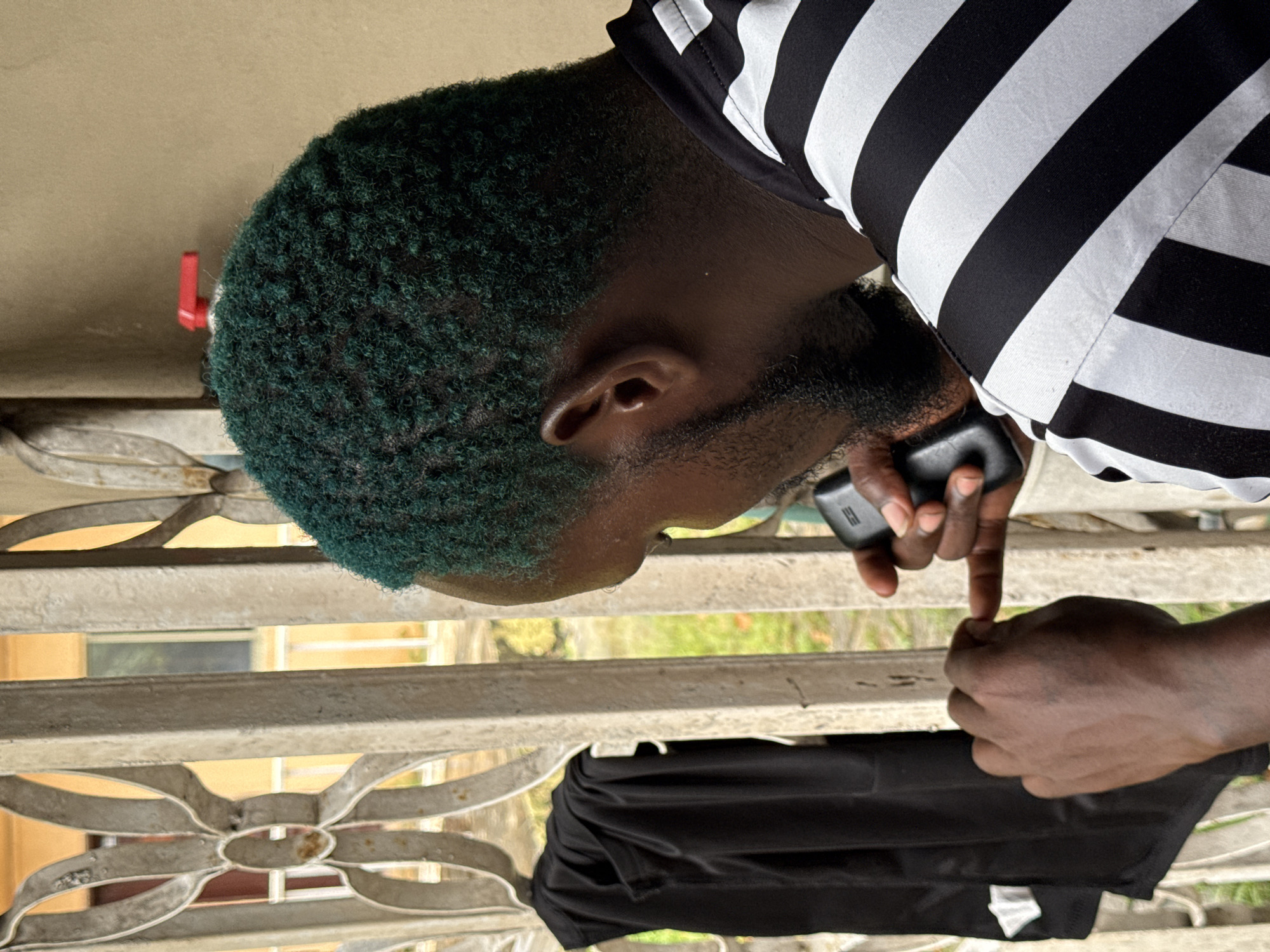 A man with dyed green hair is seated on a porch, holding a black mobile phone to his ear with his right hand. His left hand is raised, with his index finger pointing towards the phone. He is wearing a black and white striped shirt. A black garment, possibly a shirt or jersey, hangs on a metal railing to his left. The background reveals a beige wall and some greenery. This scene is likely located in Ado, Nigeria.