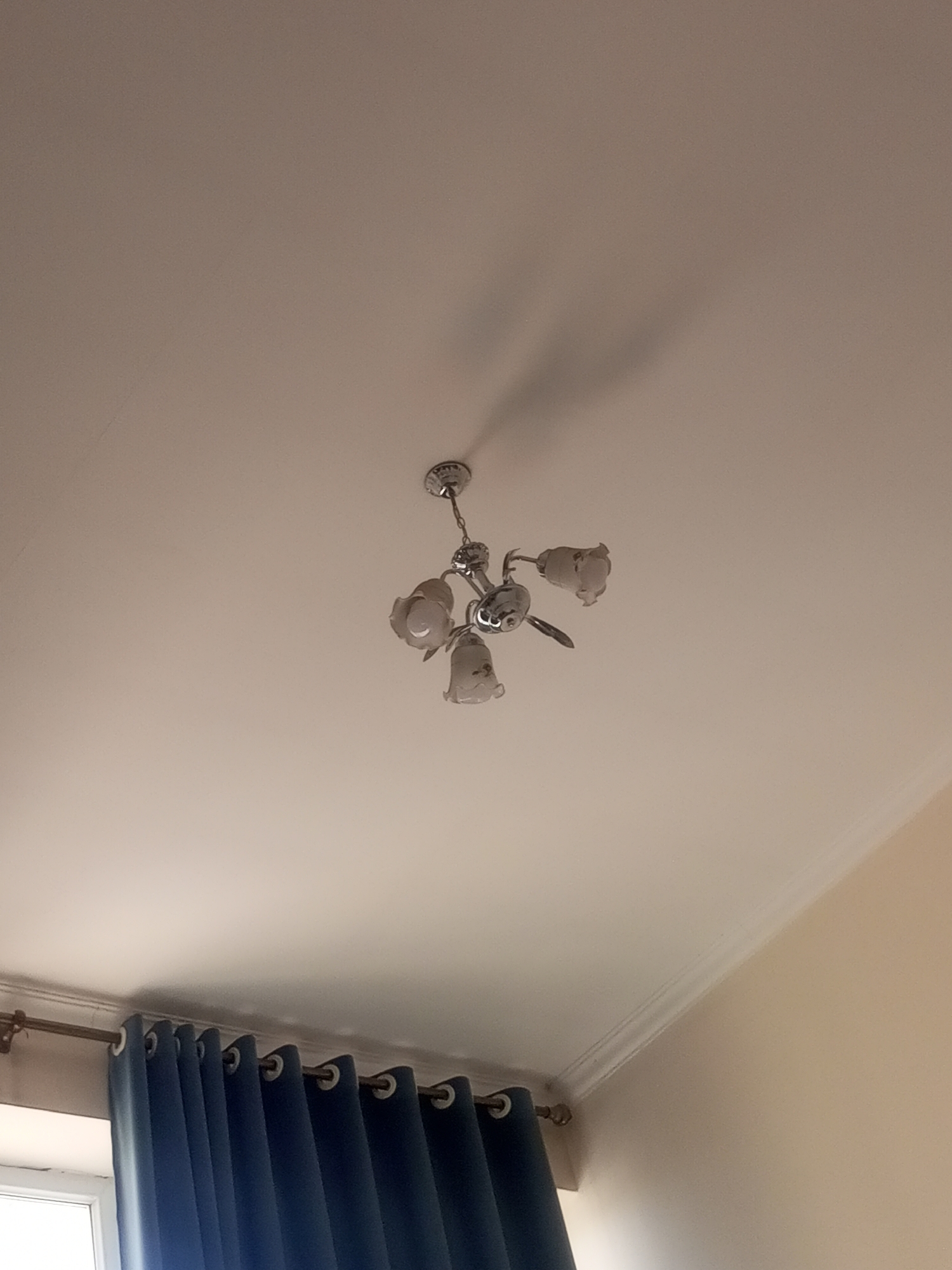 An indoor, eye-level shot shows a ceiling with a chandelier and blue curtains. The ceiling is white and has molding around the edges. A chrome chandelier with four tulip-shaped glass shades hangs from the ceiling. The chandelier is off. Blue curtains are drawn across a window, obscuring the view outside. The curtains are gathered and appear to be made of a heavy fabric. A window frame is visible behind the curtains on the left side of the image. The overall impression is that of a domestic interior. The image is undated and the location is unknown.