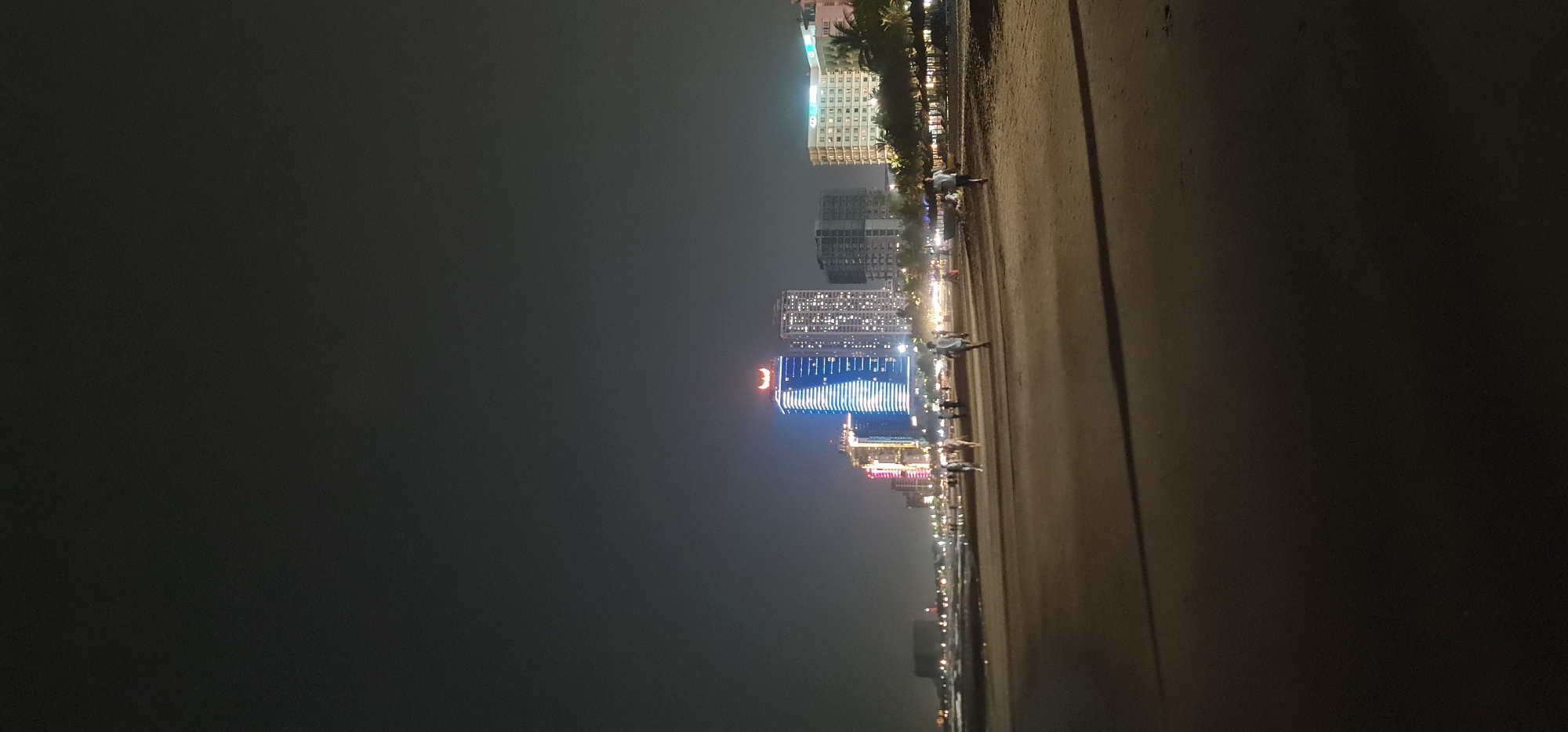 The photograph depicts a beach at night in Đà Nẵng, Vietnam. Several illuminated high-rise buildings line the coast. Pedestrians are visible on the beach. The sky is dark, suggesting night or a heavily overcast evening. The beach sand is visible in the foreground.
