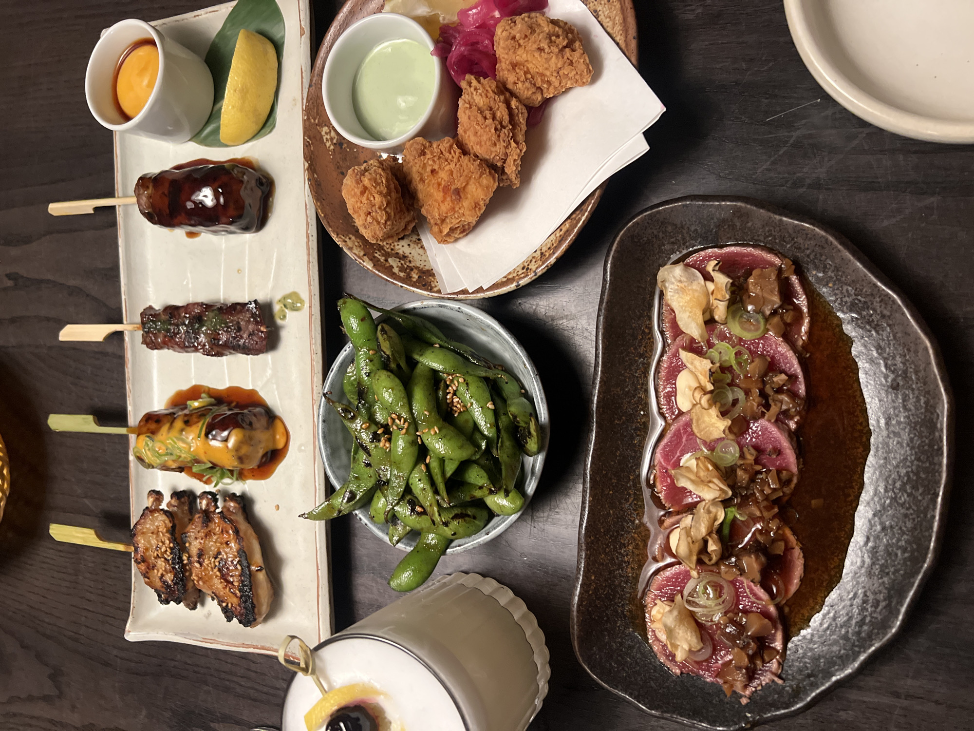 The image shows a table in a restaurant setting, laden with various dishes of Asian-inspired cuisine. On the left side of the frame, a rectangular white platter holds four different types of skewered food, accompanied by a small cup of yellow sauce and a wedge of lemon. Adjacent to it is a small bowl filled with edamame. Moving right, a brown plate contains pieces of fried chicken, accompanied by a small bowl of green sauce and some pickled vegetables. On the right, an oblong dark plate features slices of raw meat adorned with toppings. A cocktail glass sits near the bottom of the image. A single white plate can be seen in the background. The lighting is subdued. It is likely the photograph was taken at a restaurant in London, United Kingdom.
