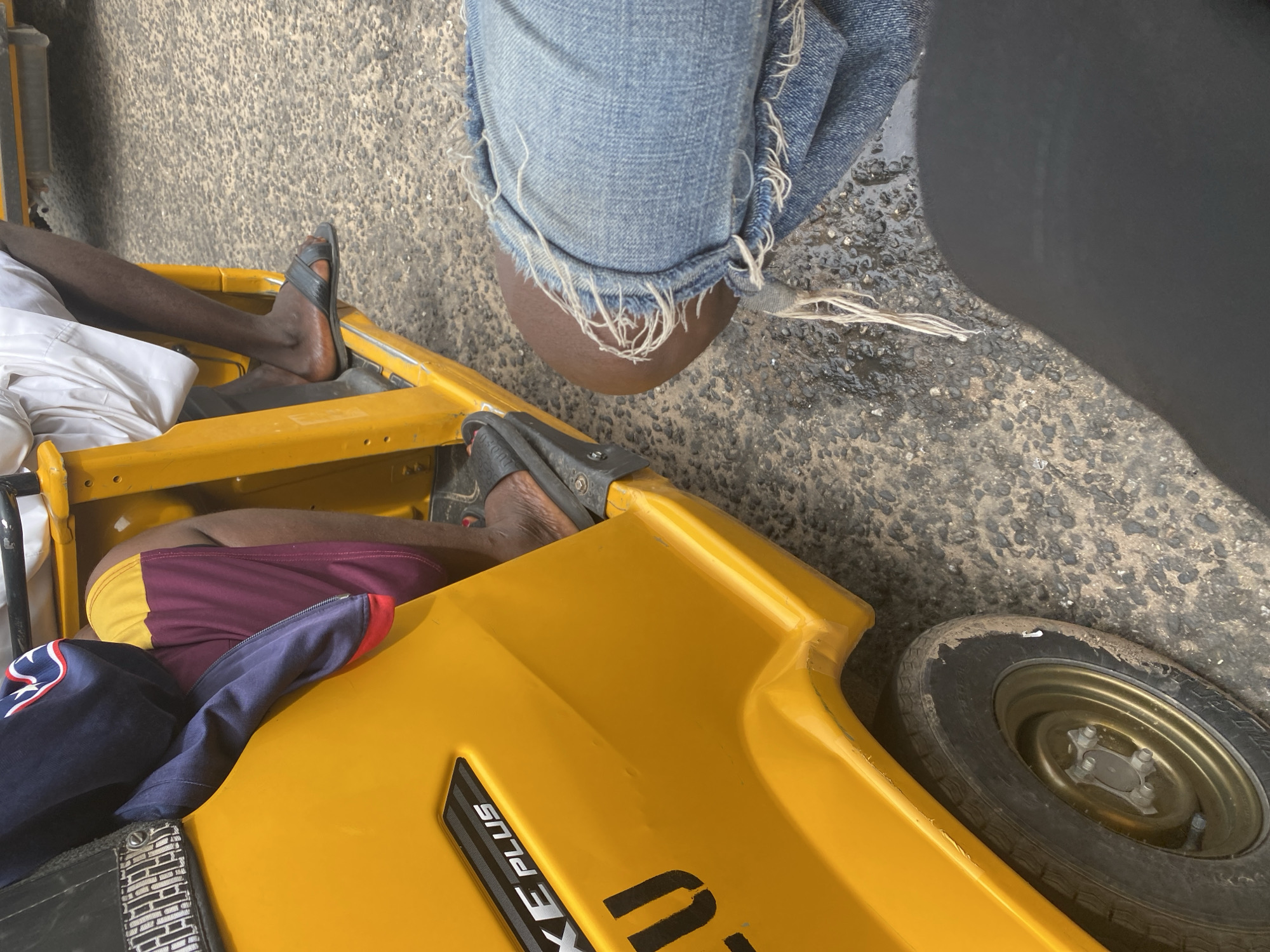 In Jalingo, Nigeria, a yellow auto-rickshaw, likely a Bajaj RE, is captured from a low-angle, close-up perspective. The vehicle's vibrant yellow exterior dominates the foreground, with visible lettering on its side reading "XE PLUS" and "LU". A portion of the front wheel, with a gold rim, is also visible on the lower left.
Inside the auto-rickshaw, two individuals are partially visible. One person, on the left, wears a dark blue jacket with red and white trim, draped over their legs which are clad in maroon and yellow shorts. Another passenger, on the right, wears a white garment and dark sandals, their leg extending towards the road.
In the foreground, to the right of the auto-rickshaw, the lower body of another person is visible, wearing ripped blue jeans. The texture of the asphalt road, wet in some areas, is discernible between the vehicle and the jeans. The overall scene suggests a moment captured during transportation within Jalingo.