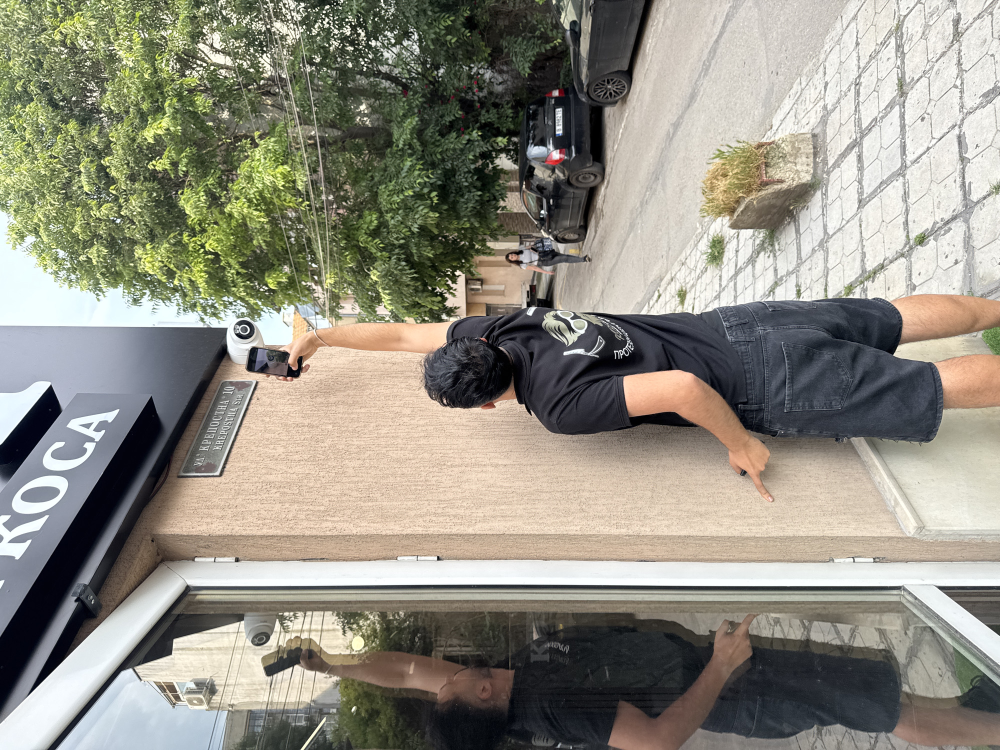 This daytime outdoor photograph captures a street scene in Varna, Bulgaria, specifically on Krēpostna Street, as indicated by a plaque reading "УЛ. КРЕПОСТНА 10 KRĒPOSTNA Str." The main subject is a man, viewed from behind, wearing a black t-shirt with a partially visible graphic including the Cyrillic text "ПРОТЕ" and black shorts. He stands in front of a textured beige building facade, holding a black smartphone aloft in his extended right hand, seemingly taking a photo or video. His left index finger points downwards towards the ground.
Above the man's position, a white security camera is mounted on the building. Below him, a large window prominently reflects his full figure and the surrounding street. To his left, a black sign with white letters "KOCA" is partially visible on the building.
The background reveals an urban setting with a paved sidewalk featuring a hexagonal pattern. Two black cars are parked along the street; one has a Bulgarian license plate "B 8492 KE," confirming Varna registration. A woman in a light-colored top and dark pants is walking on the sidewalk further down the street. Lush green trees line the street, and some overhead wires are visible through the foliage. A concrete planter with dry grass sits on the sidewalk. The bright lighting suggests a clear or lightly overcast day.