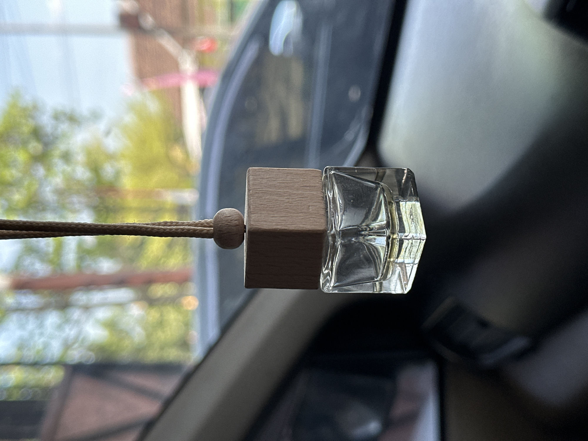 A car air freshener, featuring a clear glass bottle with a wooden cap and a small wooden bead, hangs from a string inside a vehicle. The air freshener is positioned in the foreground, with the interior of the car, including the dashboard, visible in the lower portion of the frame. Through the car's windows, a blurred outdoor scene is depicted, showing trees, buildings with red-tiled roofs, and a clear sky, suggesting an urban or suburban environment. The overall lighting indicates it is daytime. The specific location of Bulakrejo, Indonesia is not visually identifiable in this image.