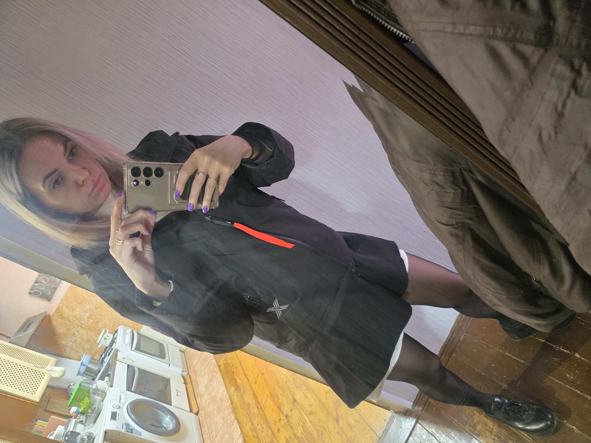A young woman is taking a selfie in front of a mirror. She is wearing a black jacket and black tights with black shoes. Her fingernails are painted purple. The background shows a kitchen with a washing machine and other appliances. A brown coat is reflected in the mirror alongside the woman. The location is implied to be in the city of Chaikovsky, Russia, based on the prompt instructions, although there is no visual evidence to confirm this.