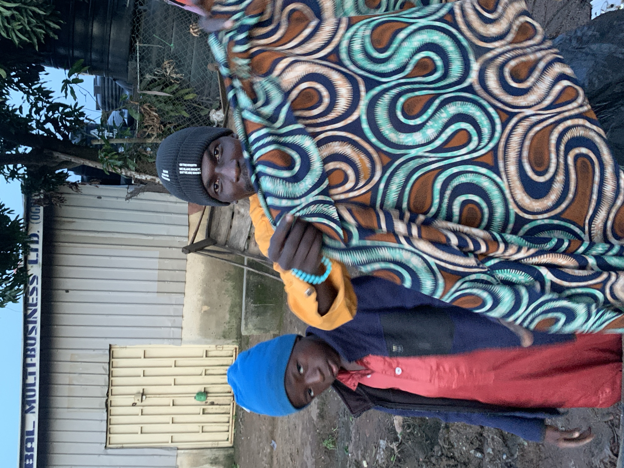 The image shows two individuals standing outside a building in Gura Top, Nigeria. A man in a black beanie and an orange shirt holds up a colorful, patterned fabric. Next to him, a young boy wearing a blue beanie and a red shirt with a dark blue jacket stands looking towards the camera. The building behind them is made of corrugated metal and has a cream-colored door. The ground appears to be dirt and there is some debris present. Trees and a water tank are visible in the background.