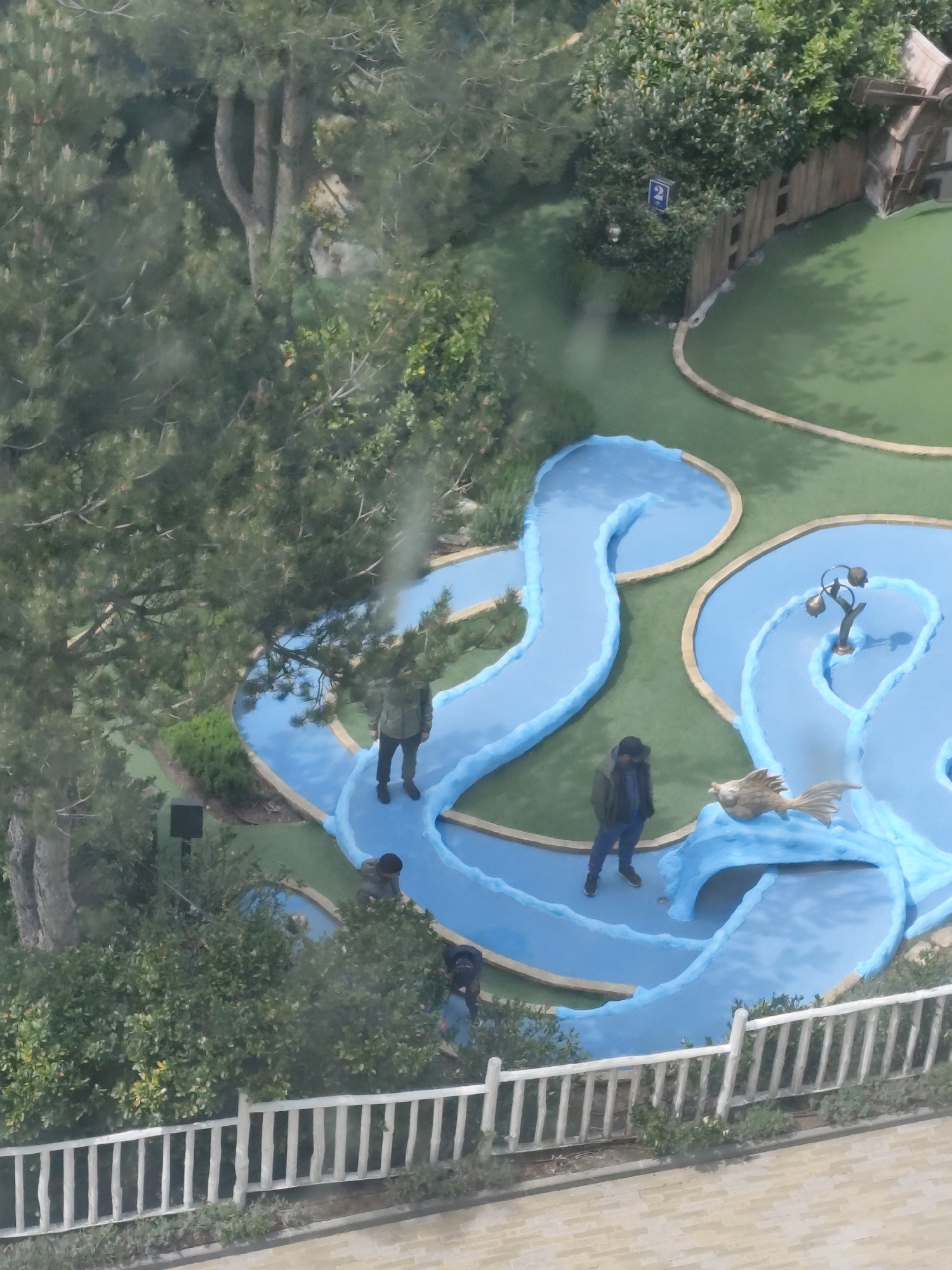 The image shows a miniature golf course. It has light-blue pathways that mimic water features, accented with white trim evoking waves. Four people are visible, all wearing casual clothing. One individual is situated further up the course, and another is near a decorative bird sculpture. Two more people are located near a white picket fence in the lower part of the frame. The golf course is lined with greenery including trees and bushes. In the upper right corner, a wooden structure with a sign is partially visible. The surrounding area appears to be outdoors, and the location is Unknown, Unknown.