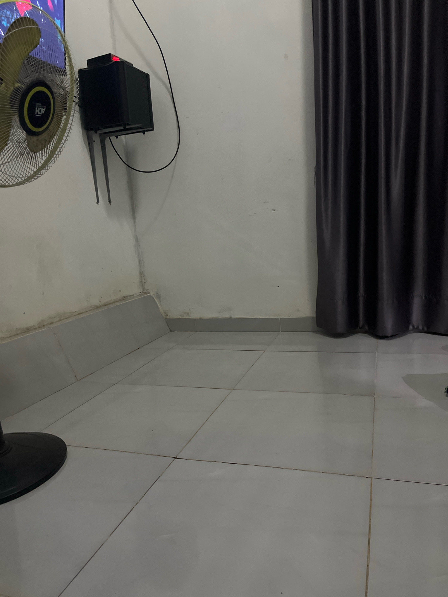 The image shows a corner of a room. In the foreground, a black fan is visible on the left, with its base on the tiled floor. The floor is covered in large, light grey square tiles with dark grout lines. The walls are painted white and show signs of some staining and dirt, particularly along the baseboards and in the corner.
Mounted on the wall is a black rectangular device with a red light illuminated on its top surface. This device is supported by two L-shaped metal brackets. A black wire extends from the device upwards and to the right. To the right of the device, a dark grey curtain hangs from the wall, covering an unseen area. The lighting in the room appears to be dim, with shadows cast by the objects present.
The scene is located in Kebumen, Indonesia.