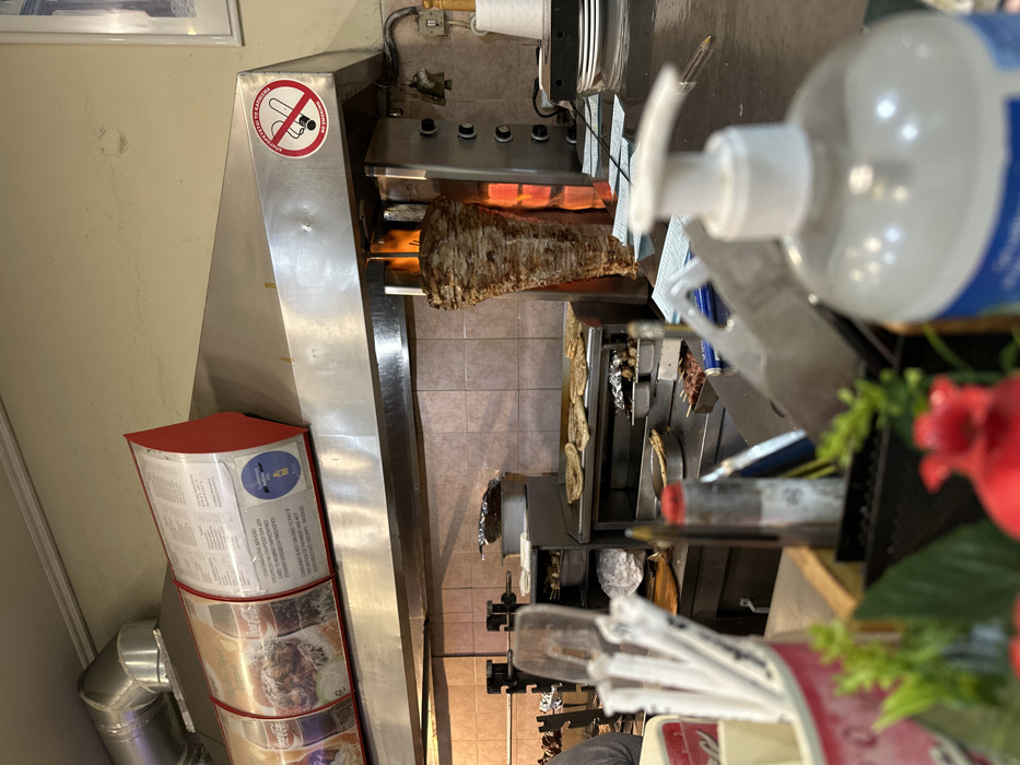 This image captures the bustling interior of a casual food establishment, likely a gyros or souvlaki shop, focusing on the food preparation area.
At the center, a large, vertical spit of seasoned meat, characteristic of gyros or shawarma, is rotating and cooking over radiant heat, its outer layer visibly browned. The cooking apparatus is made of gleaming stainless steel, with burners providing the heat. Above the spit, a stainless steel hood or ventilation system is visible.
To the left of the cooking station, a prominent circular sign is affixed to a stainless steel panel, depicting a "no smoking" symbol (a lit cigarette with a red cross through it). Below the symbol, text in Greek reads "ΑΠΑΓΟΡΕΥΕΤΑΙ ΤΟ ΚΑΠΝΙΣΜΑ" and in English, "NO SMOKING." Further to the left and lower, a red, multi-tiered display rack holds menus, some of which are adorned with "Coca-Cola" branding. An industrial ventilation pipe is also visible in this area.
In the foreground on the right, a large pump bottle of hand sanitizer is prominently displayed, slightly out of focus. Next to it, a container holds various disposable utensils, including straws and plastic forks. Some red artificial flowers and greenery provide a touch of decor.
Behind the main meat spit, a stainless steel workstation or counter holds other food items, including what appear to be skewers of meat (possibly souvlaki) and flatbreads on trays, suggesting other grilled options are prepared here. The floor is tiled, and the walls are a light, neutral color, indicating a clean and functional environment.
The scene depicts an active food service operation, common in Greece, emphasizing hygiene with the visible hand sanitizer. No people are visible in the frame, and the time of day cannot be determined.