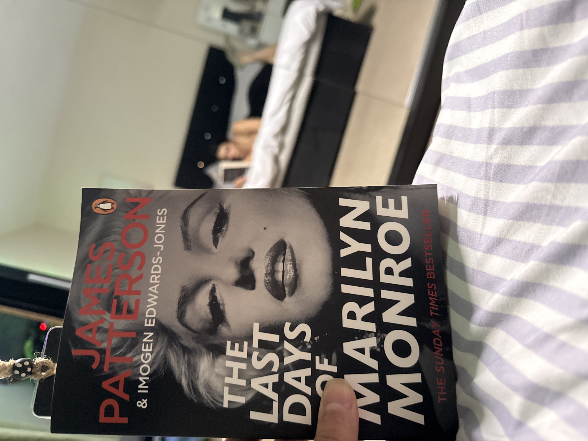 The image shows a paperback book titled "The Last Days of Marilyn Monroe" by James Patterson and Imogen Edwards-Jones. The book cover features a black-and-white portrait of Marilyn Monroe. The book is placed on a bed with striped bedding. In the background, a blurry figure of a person lying in bed and using a tablet is visible. The location, Great Yarmouth, United Kingdom, is not discernible from the image.